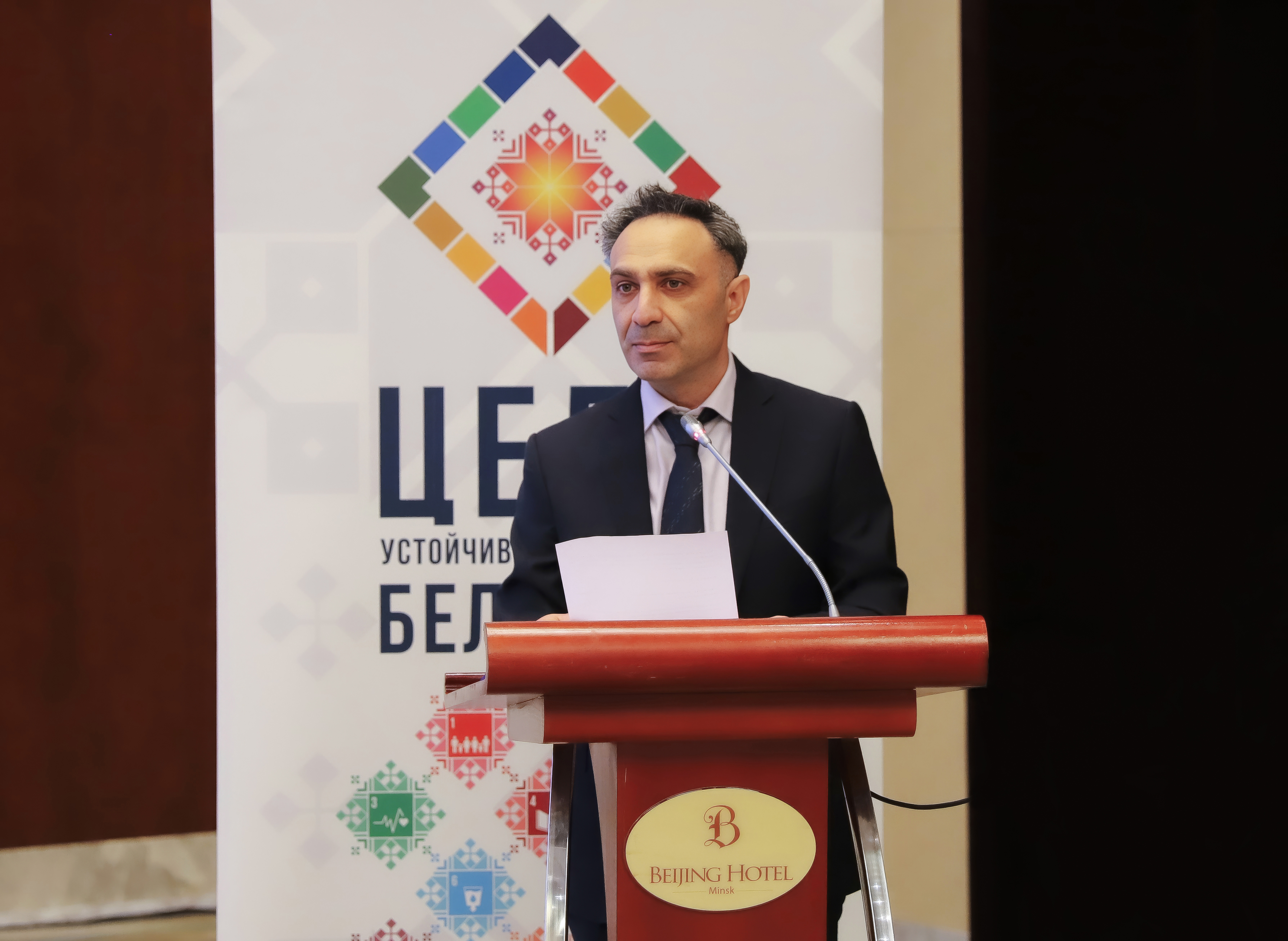 Man in suit at wooden podium, colorful geometric logo banner behind.