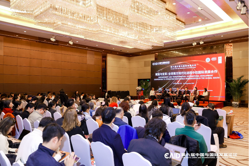 Large conference hall with crystal chandeliers; audience seated facing a stage with a red banner.