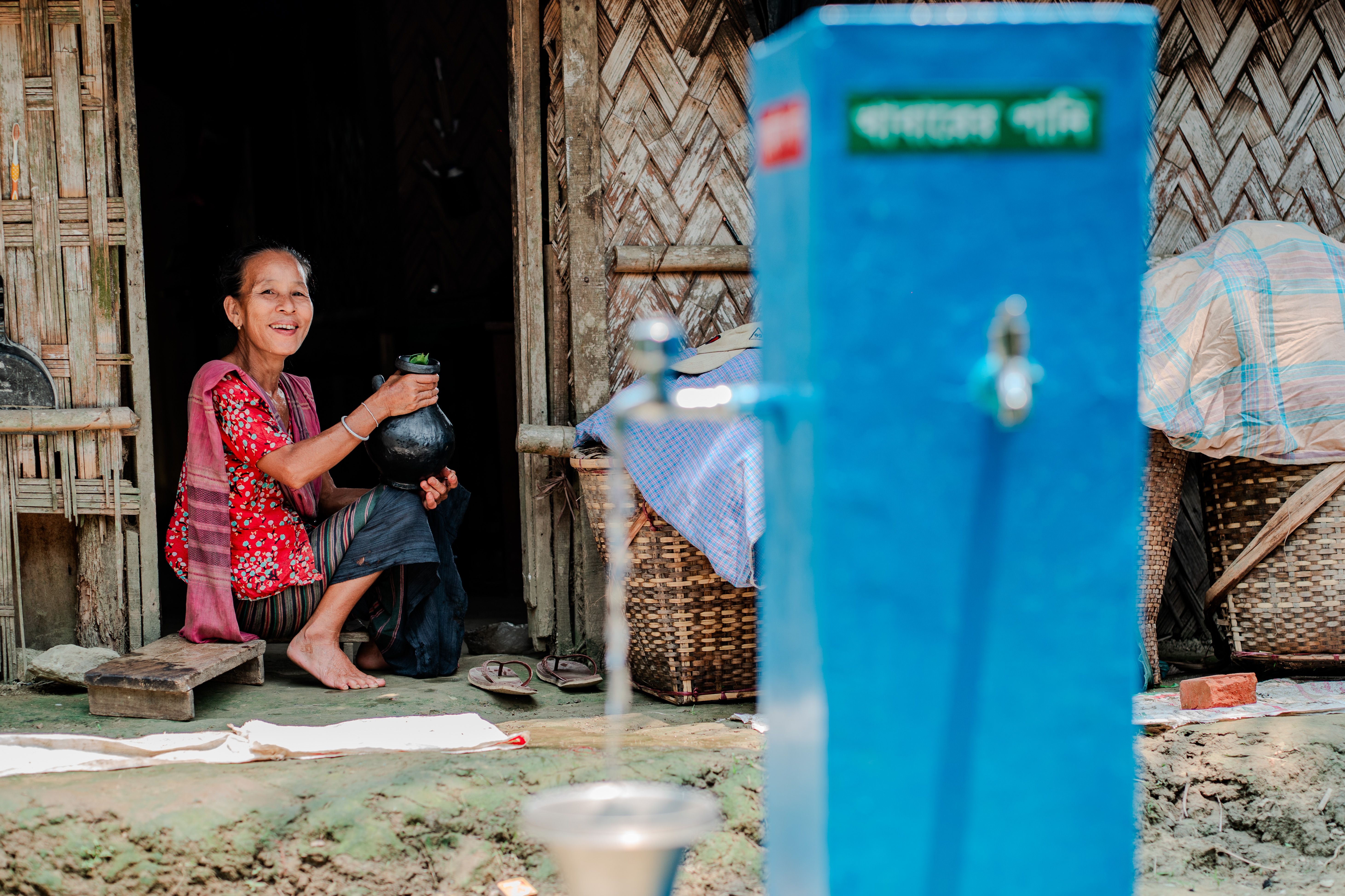 Two women sit on a wooden village porch beside a tall blue water dispenser.
