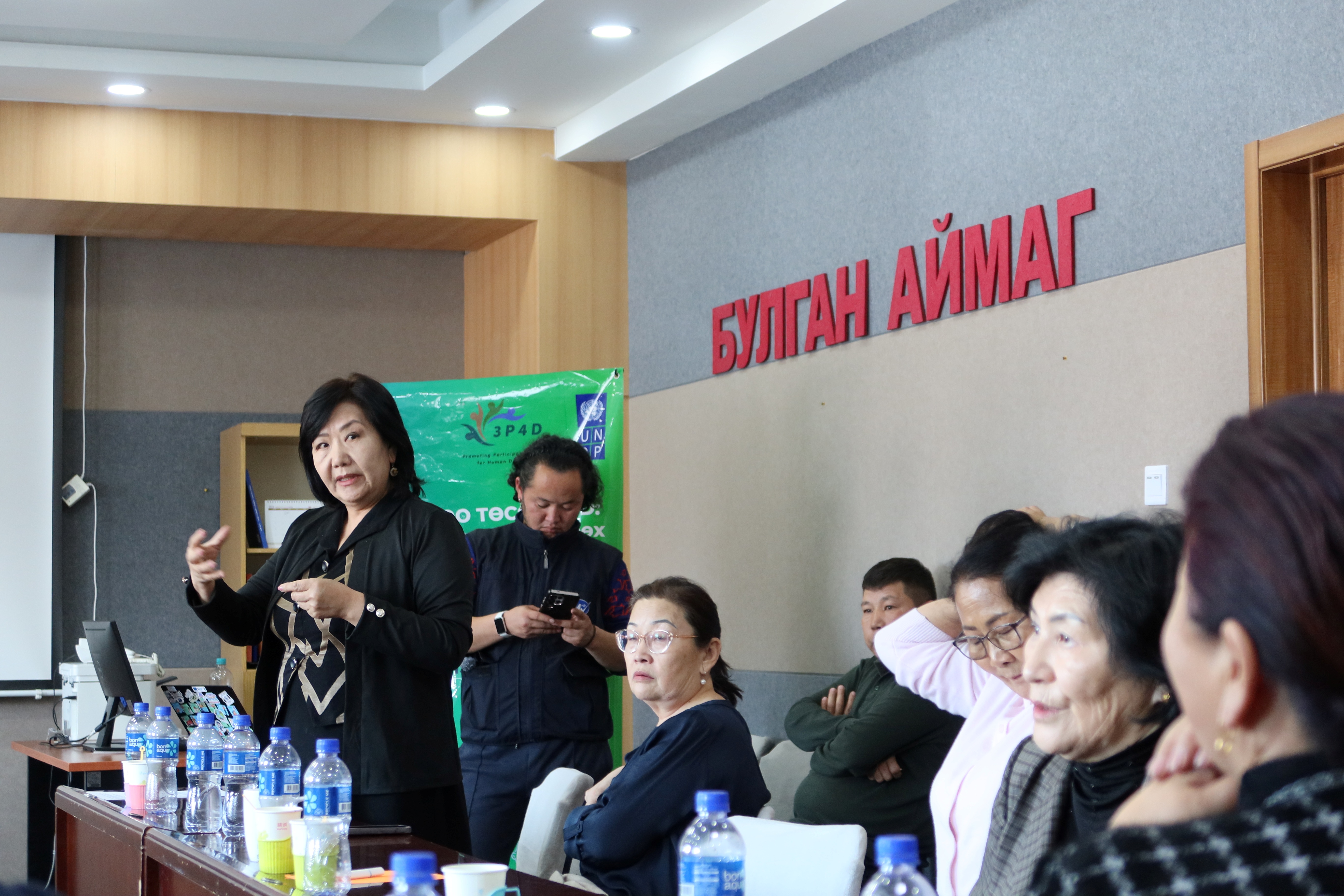 A woman speaker gestures to a seated audience in a conference room.