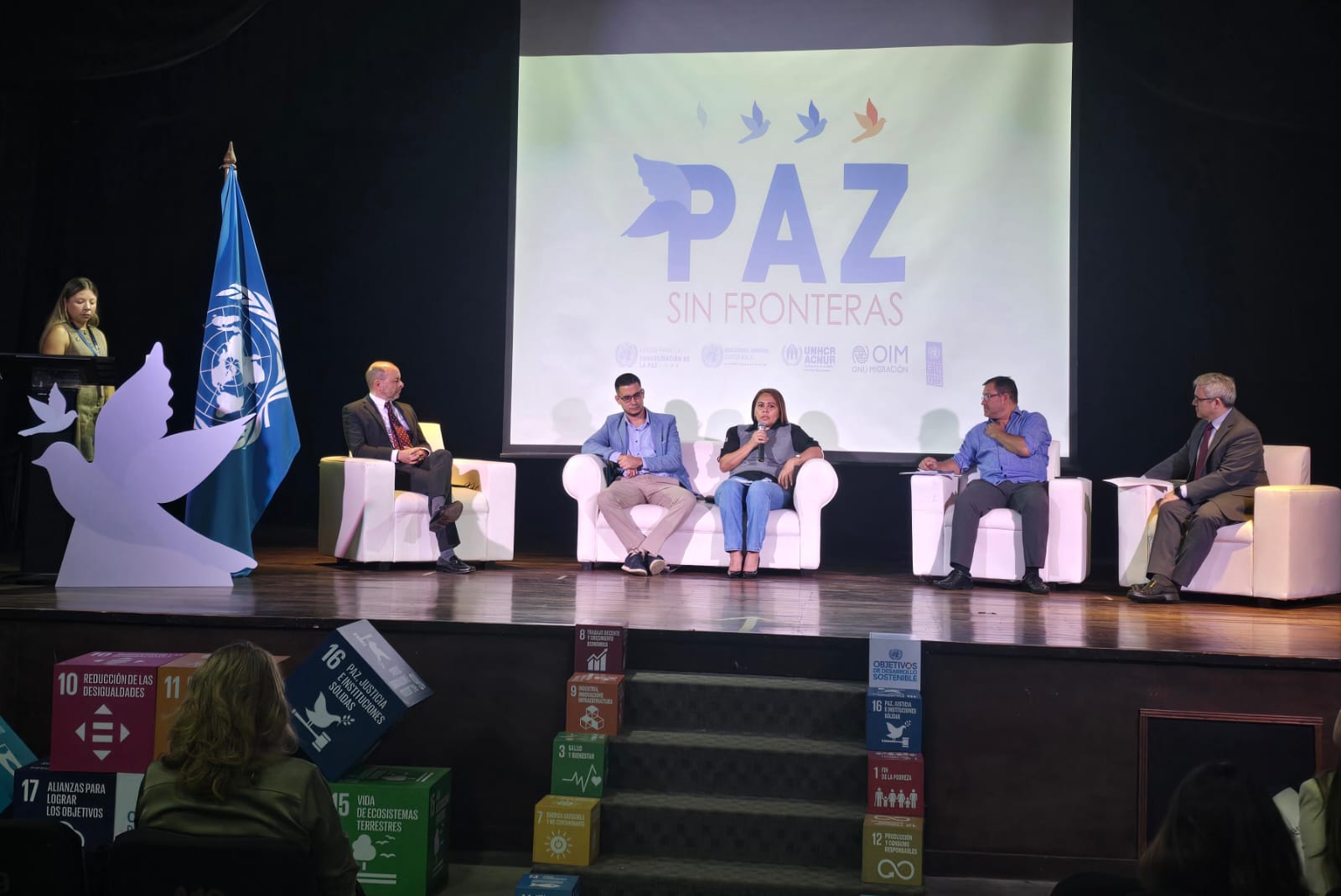 Photograph of a peace panel on stage with PAZ logo and seated panelists, flags on the left.