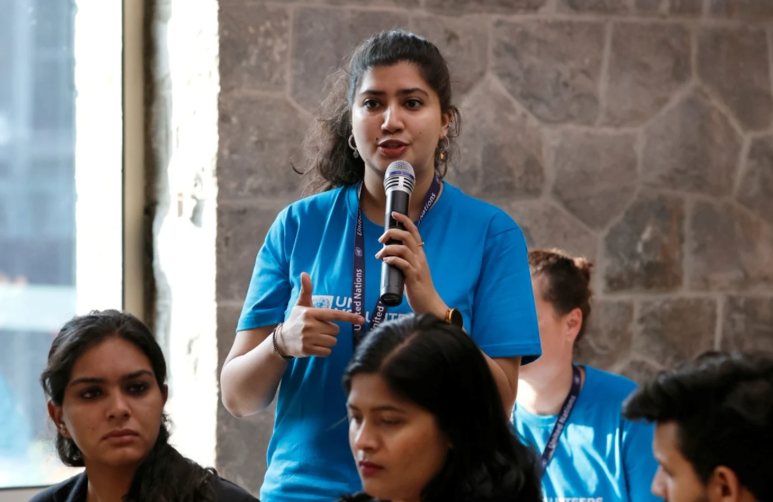 Speaker in blue shirt holds microphone at group event, with others seated and stone wall backdrop.