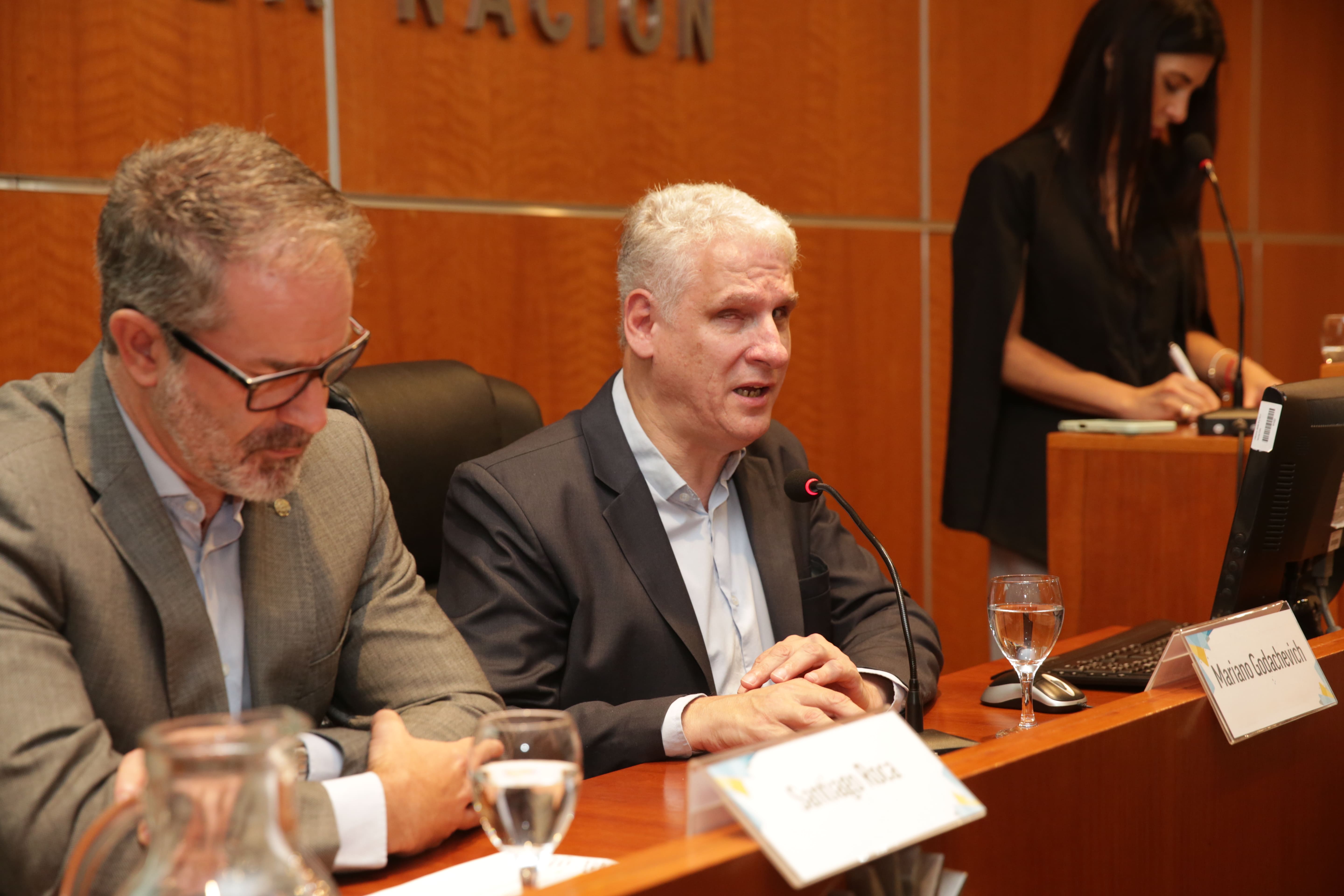 Two men in suits seated at a conference table; speaker at podium in background.