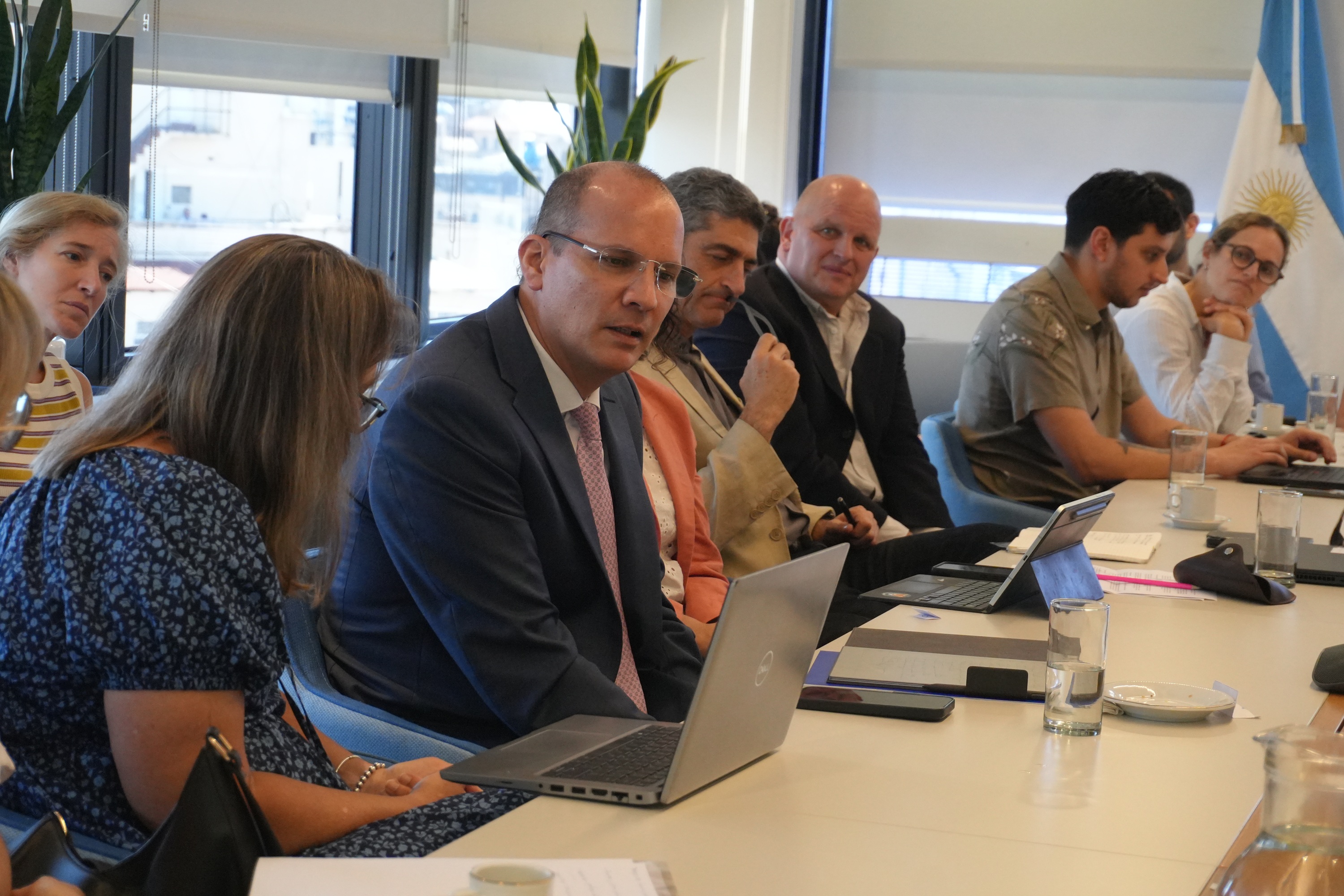 Group of professionals in a meeting around a long conference table with laptops; Ukrainian flag.