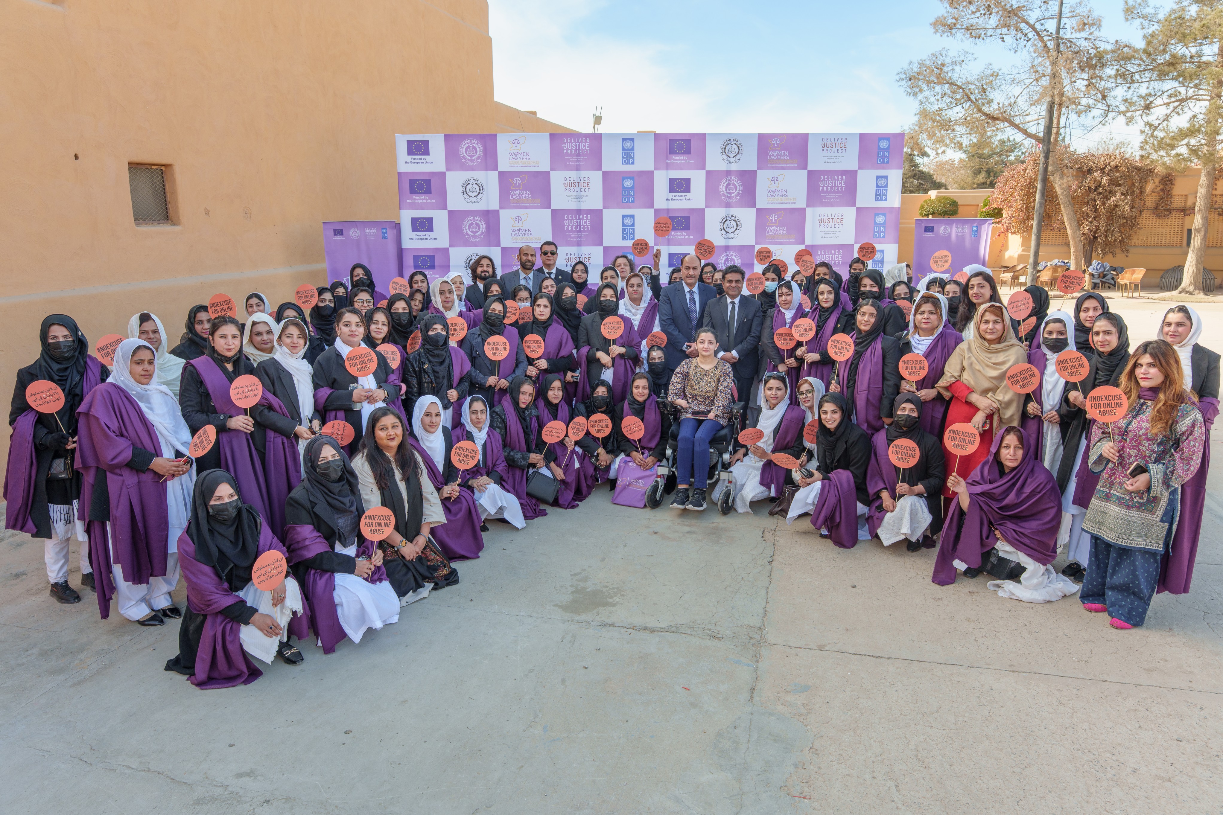 Group photo: large group in purple posing outdoors beneath a purple checkered backdrop.