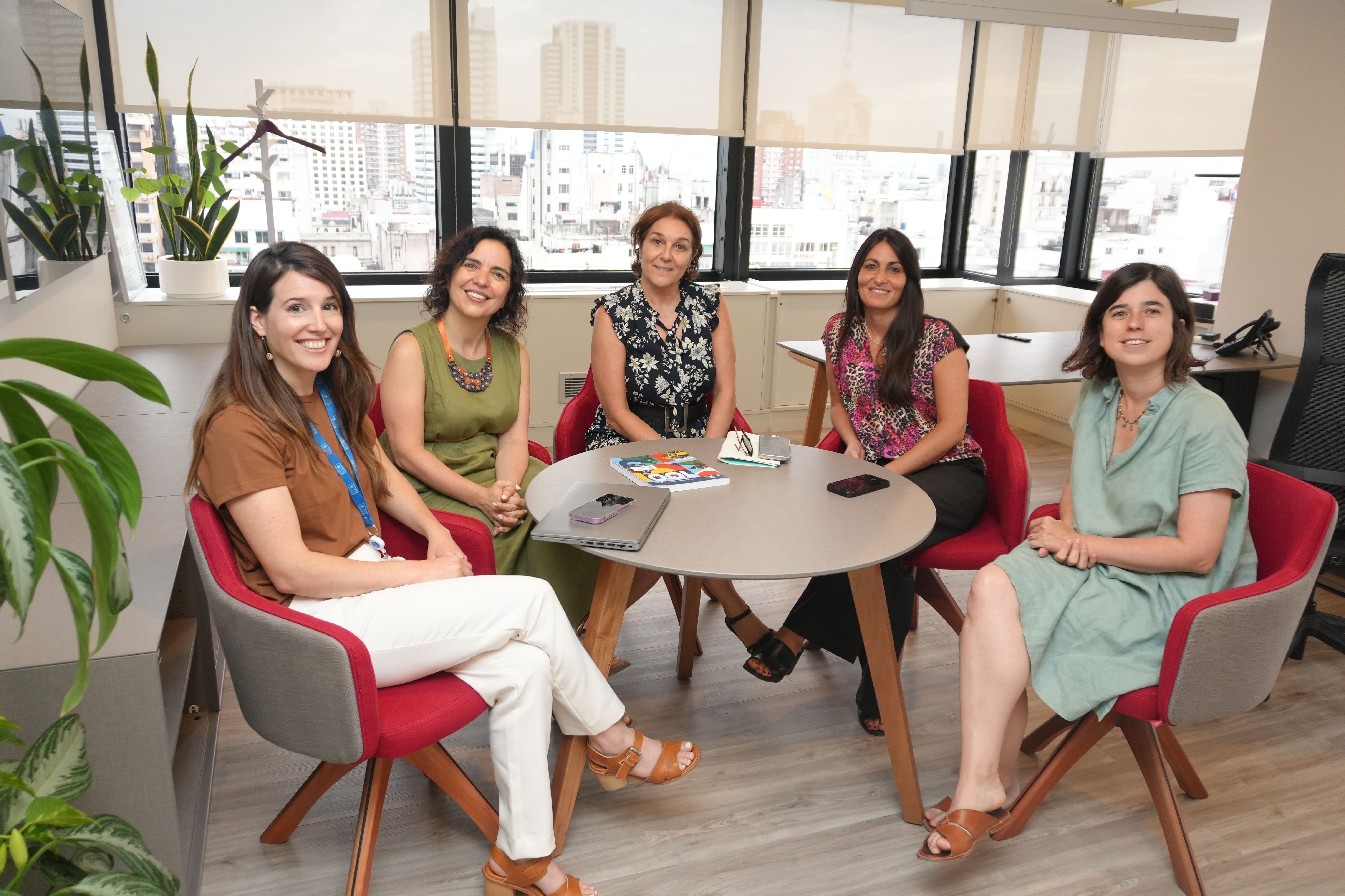 Five women sit around a round table in a bright office lounge, smiling.