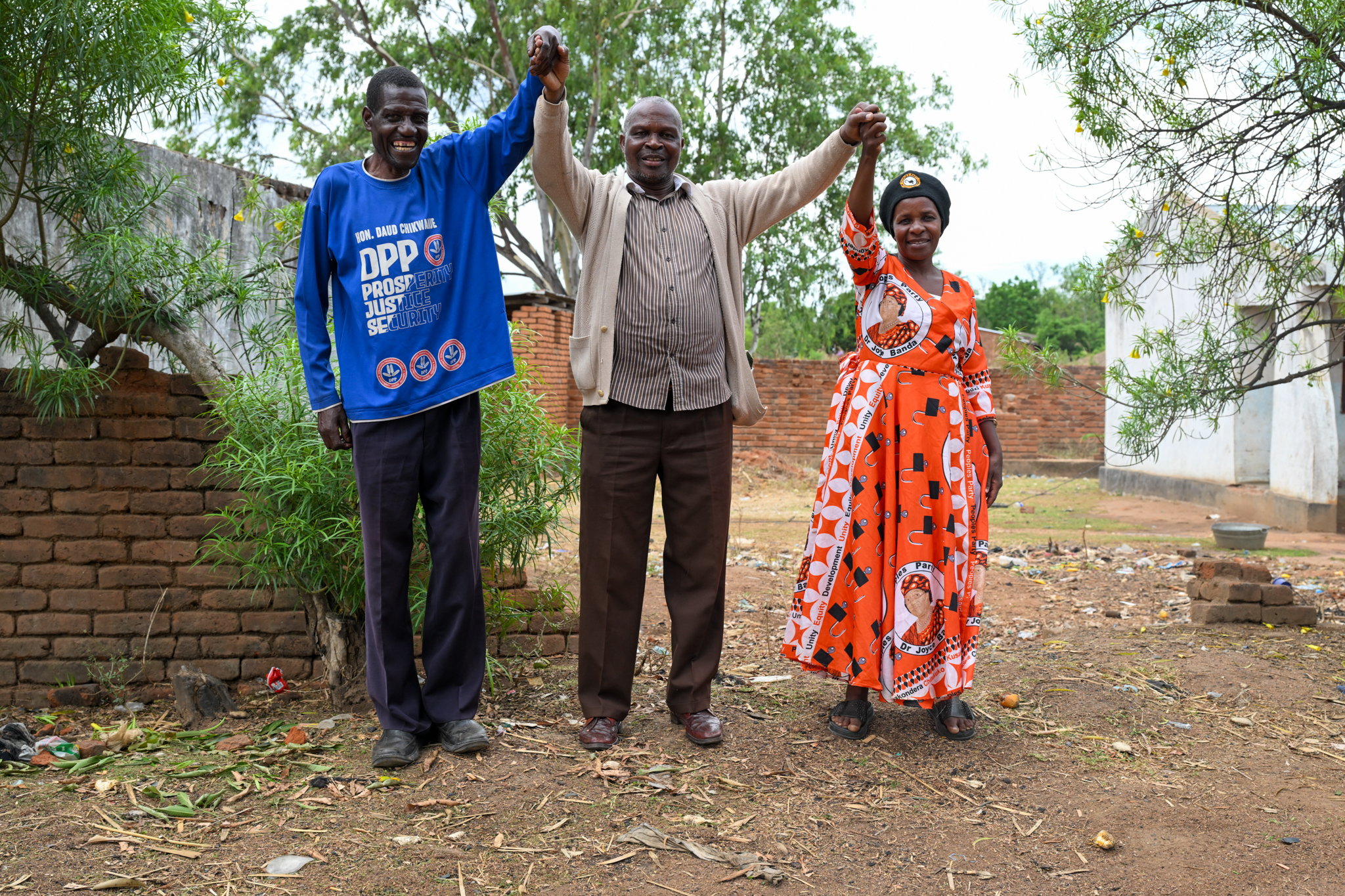 Three people outdoors, standing on dirt, raising hands in celebration; brick wall and trees behind.