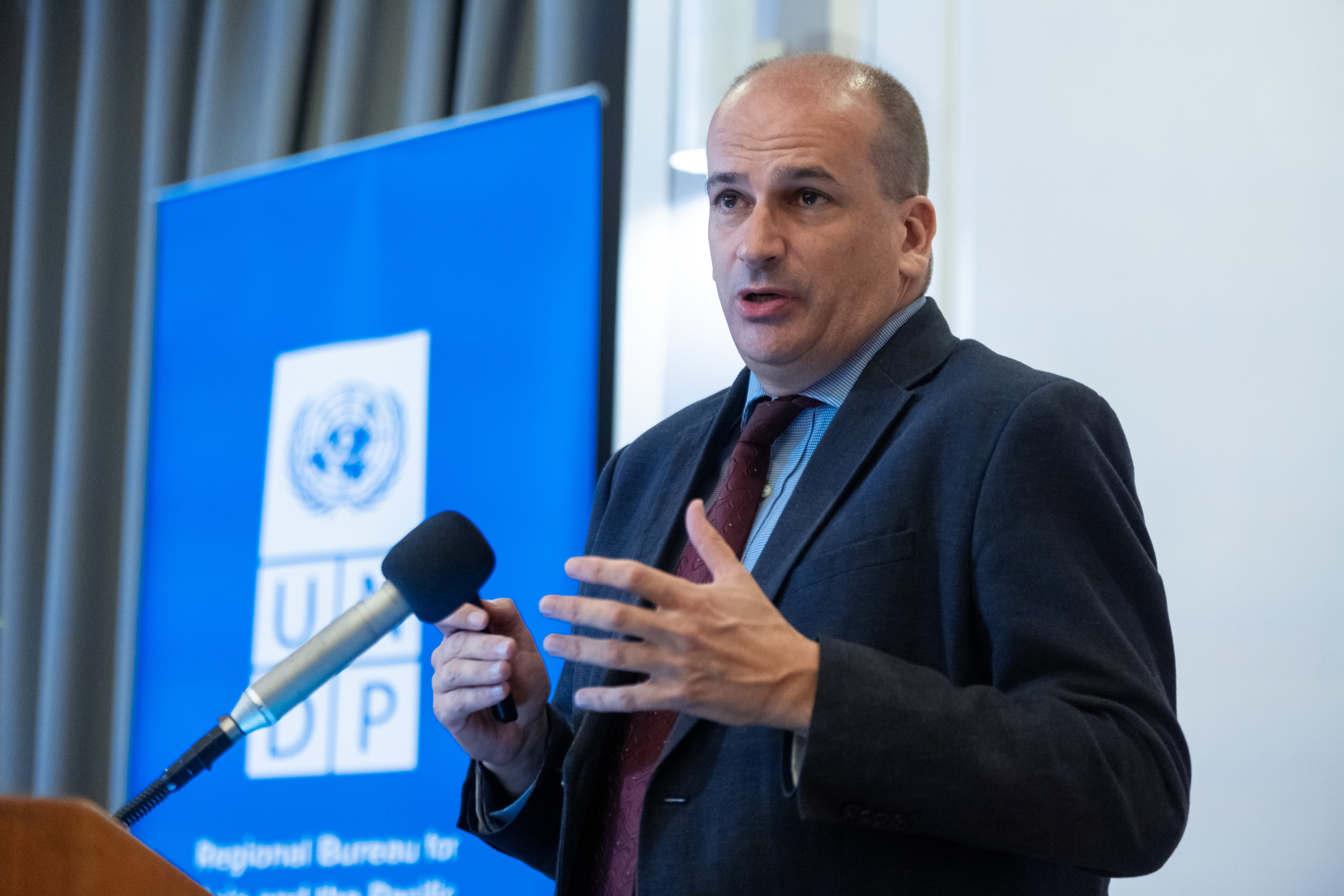 Male presenter in a dark suit and tie speaks into a microphone with a UN banner in the background.