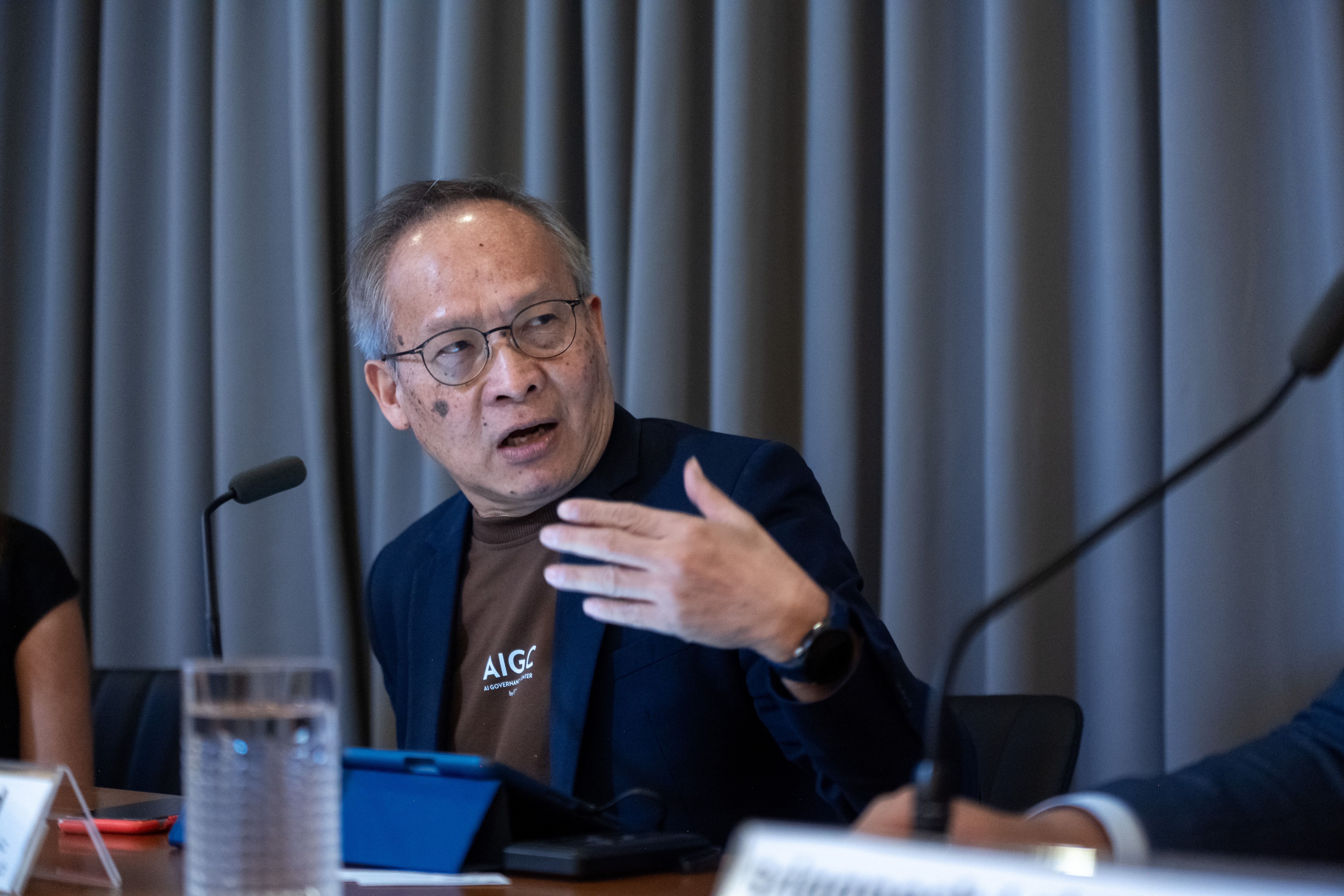 A panelist gesturing during a conference with microphones and water on a table before blue curtains.