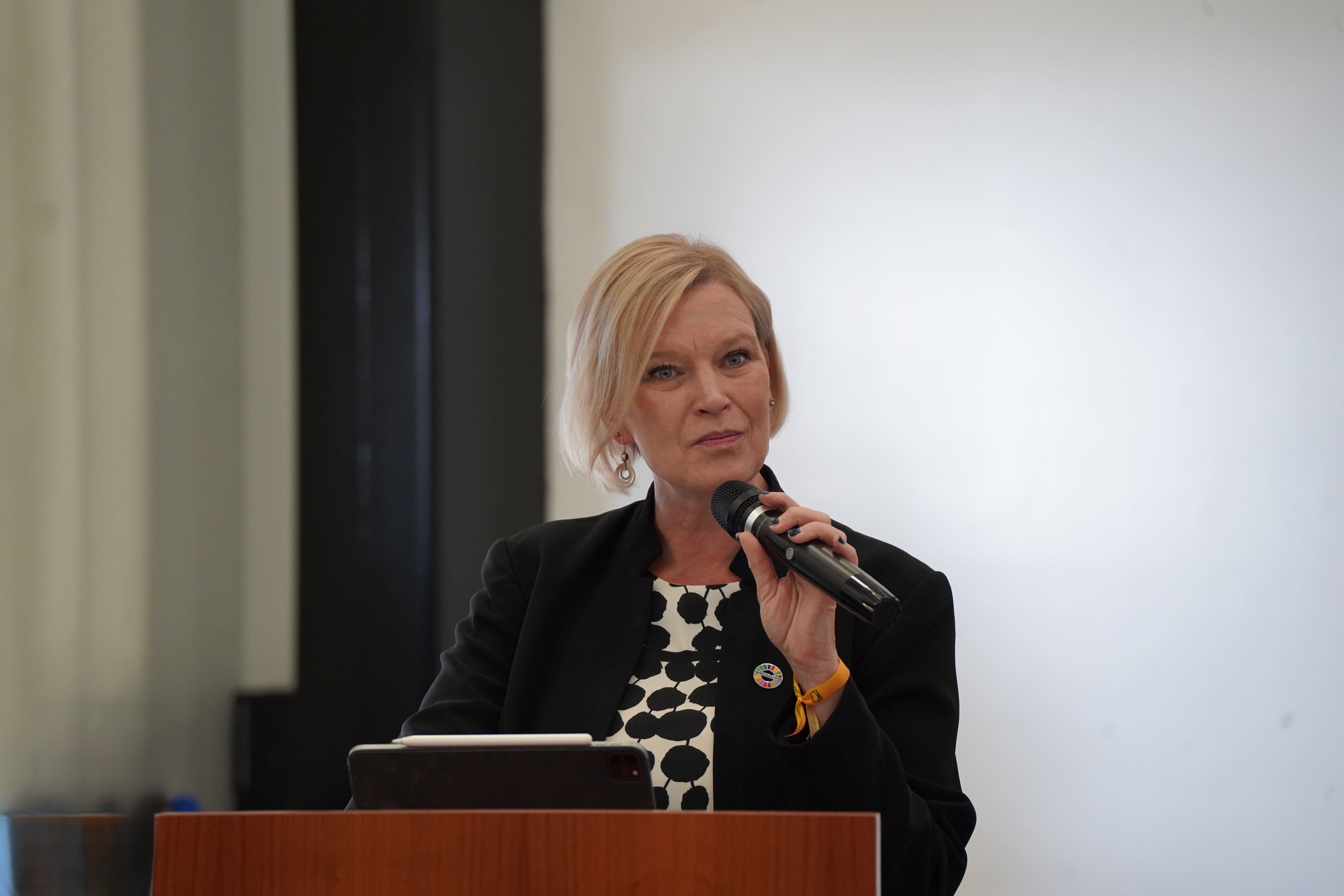 Speaker at podium presenting, microphone in hand, laptop nearby.