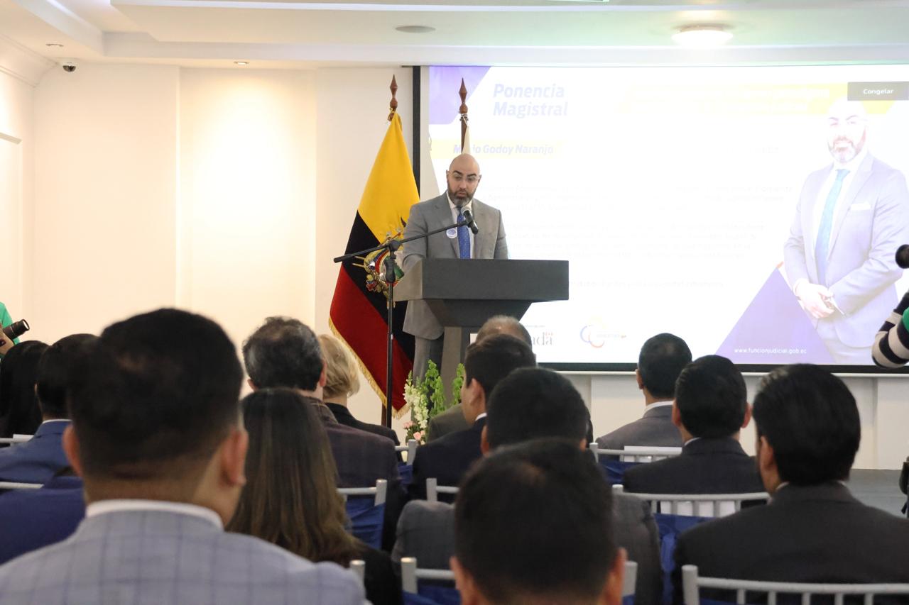 Speaker at a podium addressing an audience in a conference room with a Belgian flag.