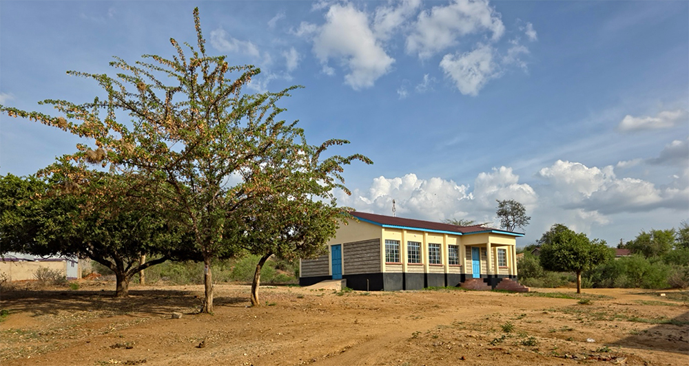 Photograph of a single-story yellow building in a dry rural area, with two trees and blue sky.