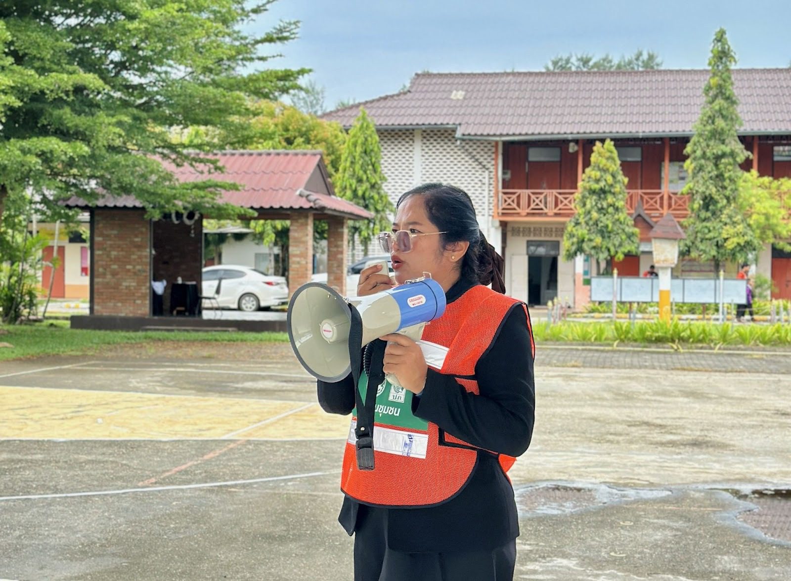 Photograph of a woman in an orange safety vest holding a megaphone outdoors on a campus.