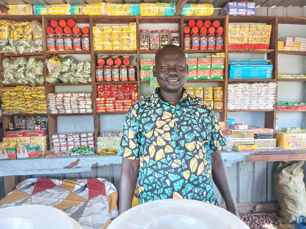 Vendor in blue patterned shirt stands behind a market stall with shelves of packaged goods.