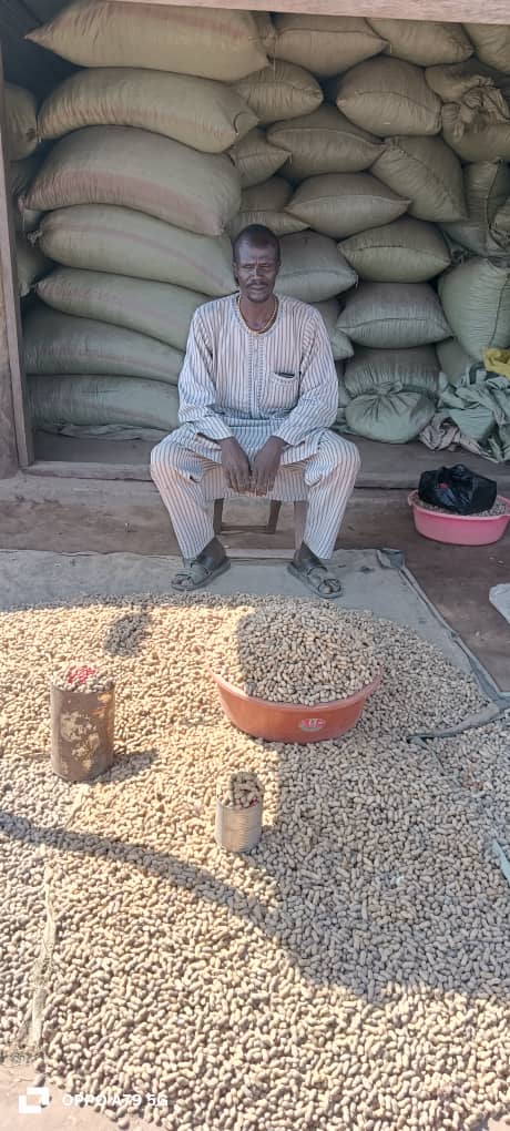 Photograph of a man seated on a chair in front of stacked burlap sacks.