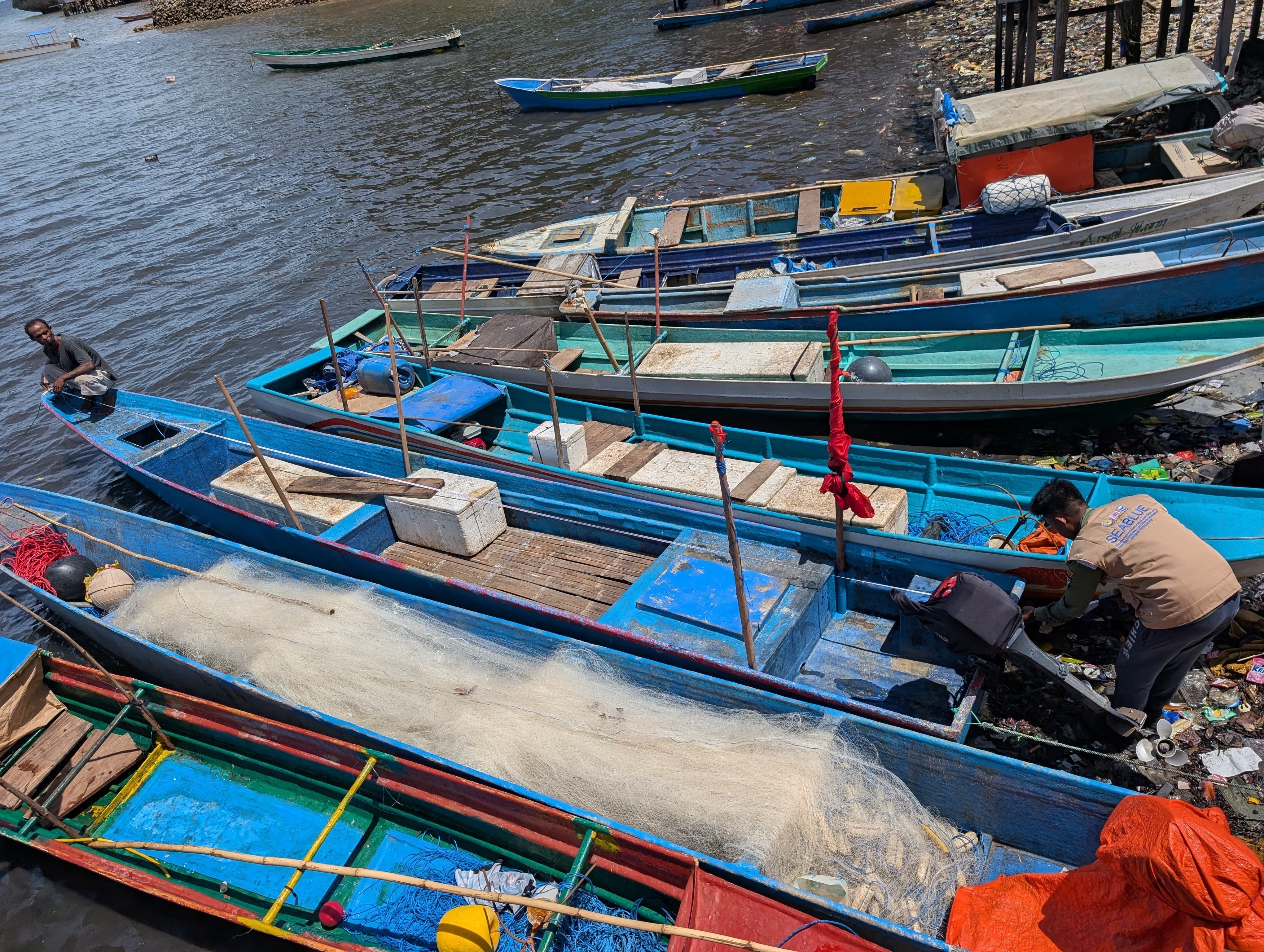 Colorful boats docked in a busy harbor; people aboard and nets scattered.