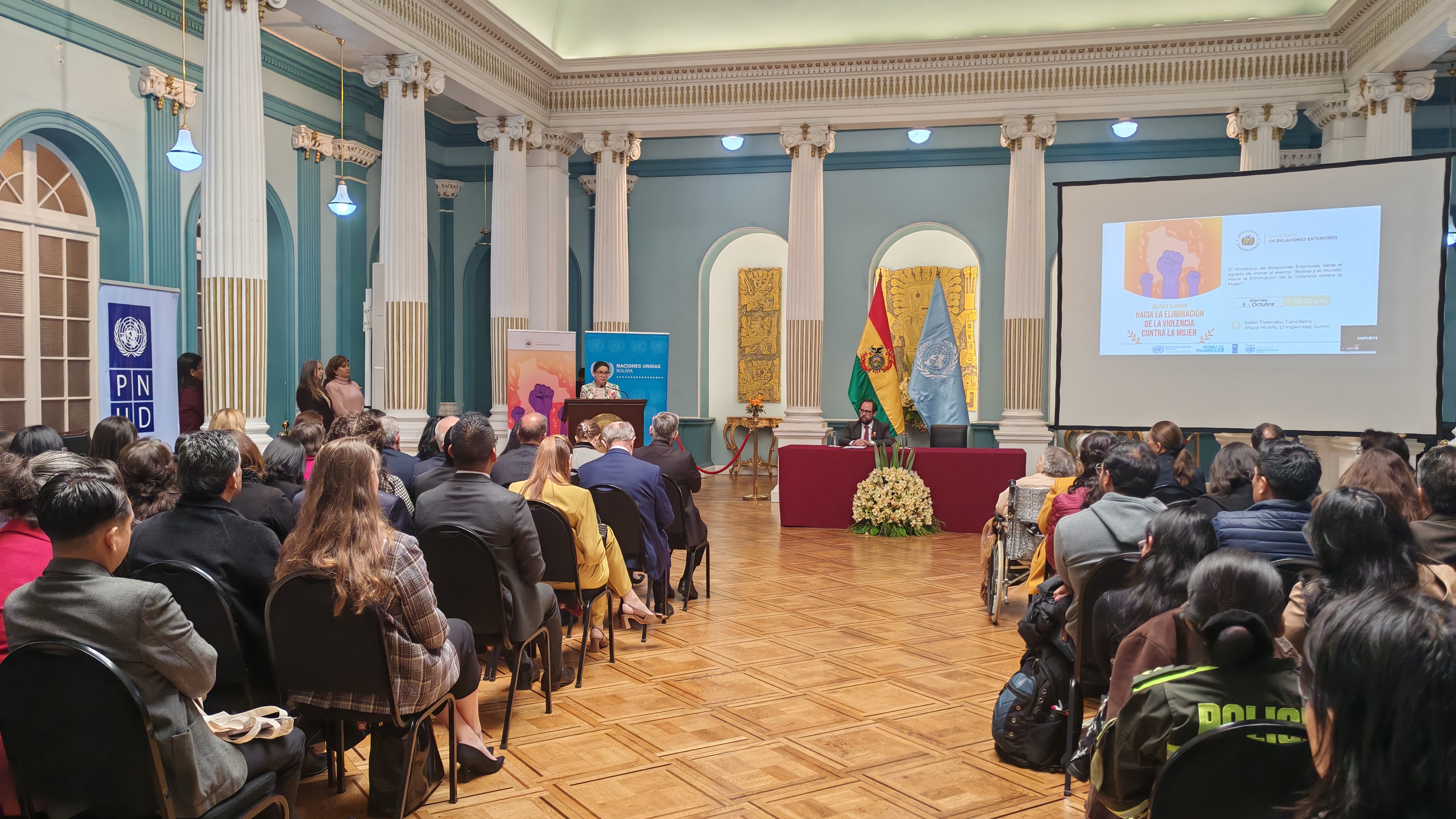Photo: ornate hall with blue walls and columns; audience watches projected presentation.