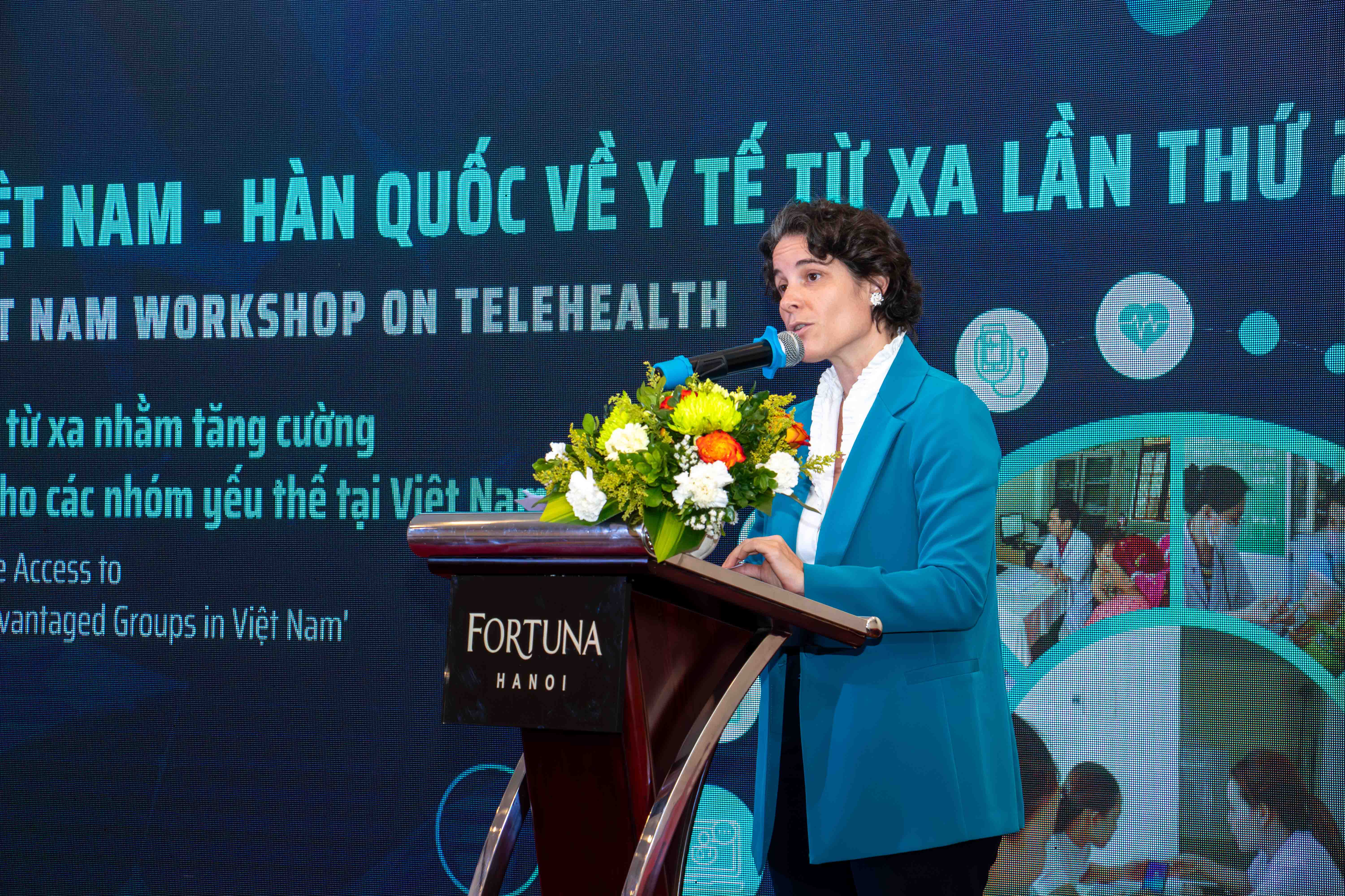 Man in blue blazer speaks at a podium on a conference stage with a teal banner in the background.