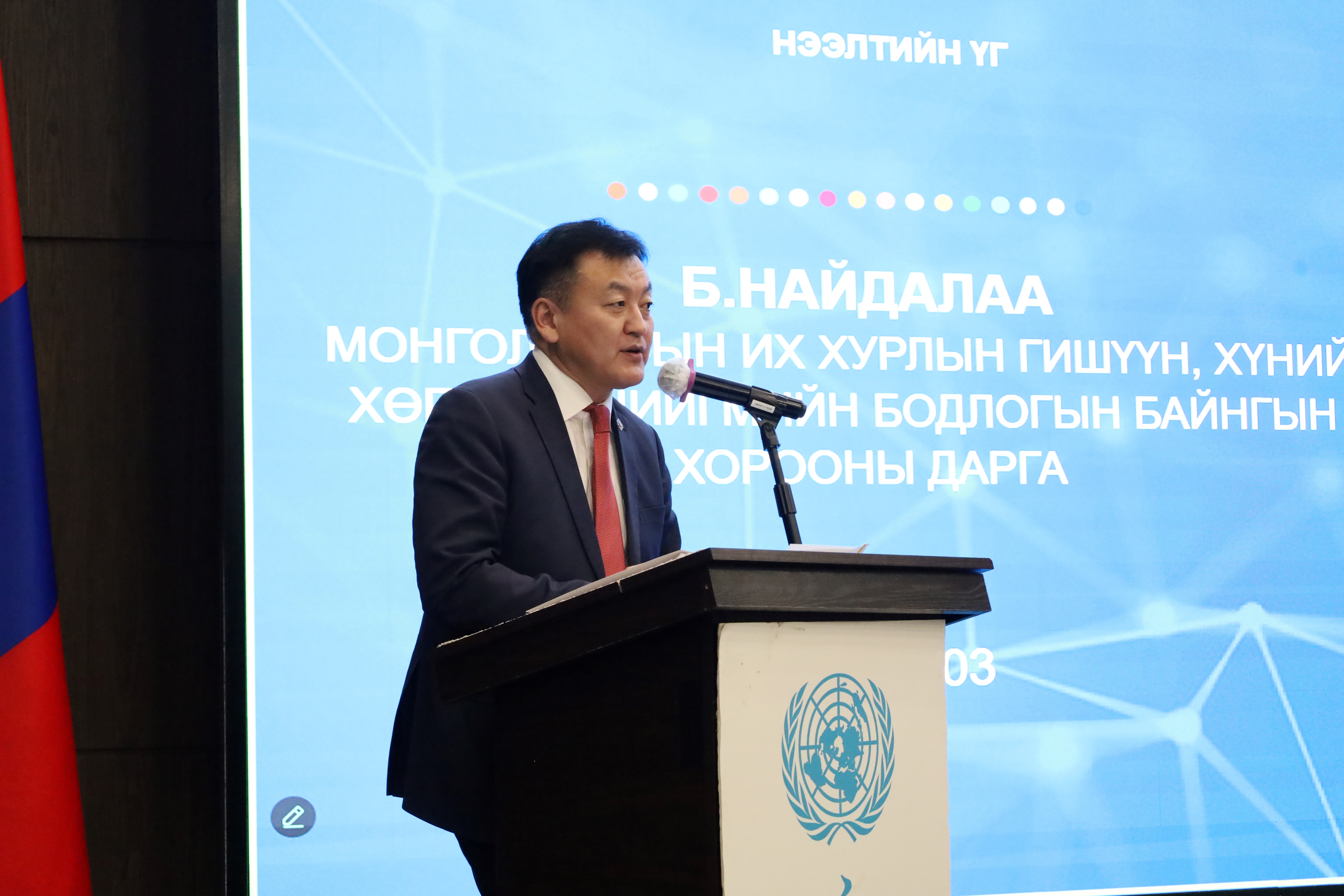 Speaker in a dark suit and red tie at a podium with a logo, blue tech backdrop, and Mongolian flag.