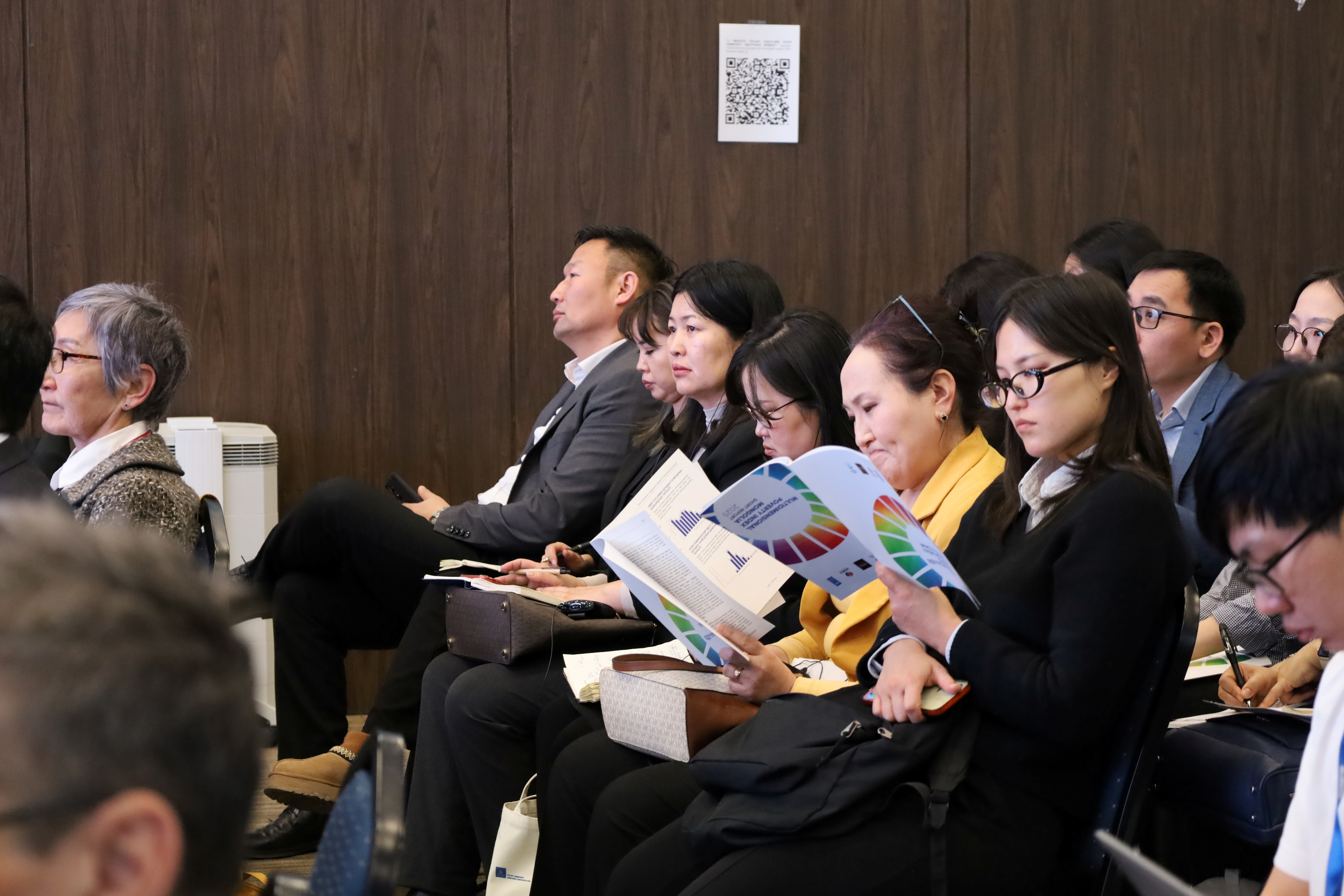 Audience seated in a conference hall; many reading colorful brochures.