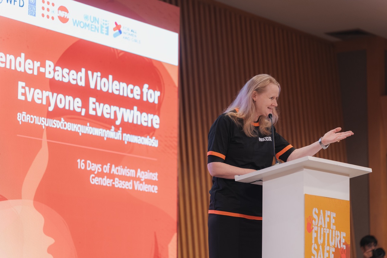 Female speaker at a podium presenting on gender-based violence; orange conference banners behind.