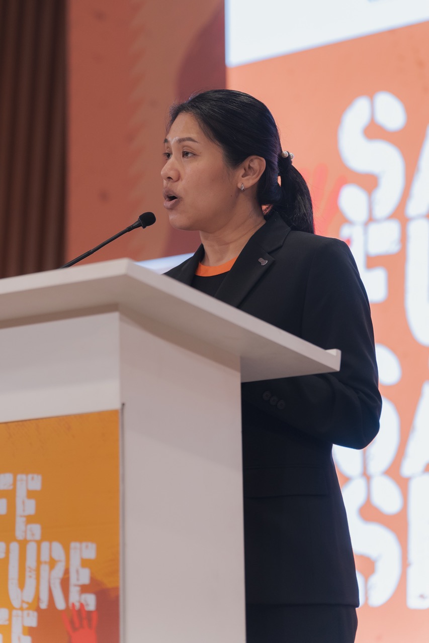 Woman speaker at podium with microphone, delivering presentation on an orange-themed stage.