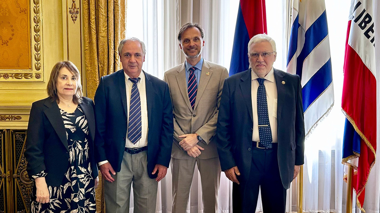 Five people in business attire stand together in a formal room with gold walls and flags.