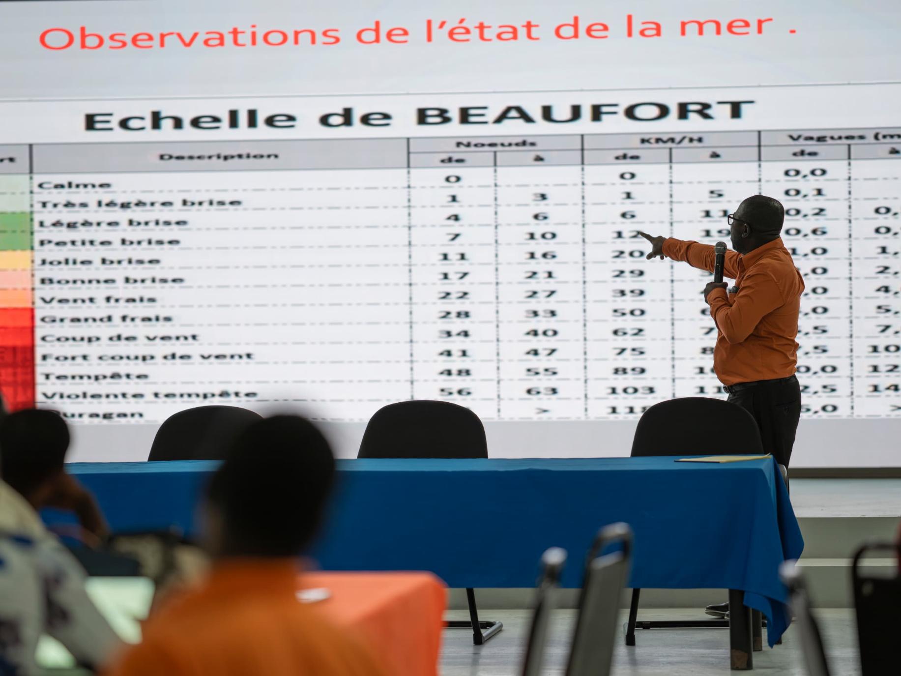 Photograph of a presenter in orange pointing at a Beaufort scale table on a large screen in a classroom.