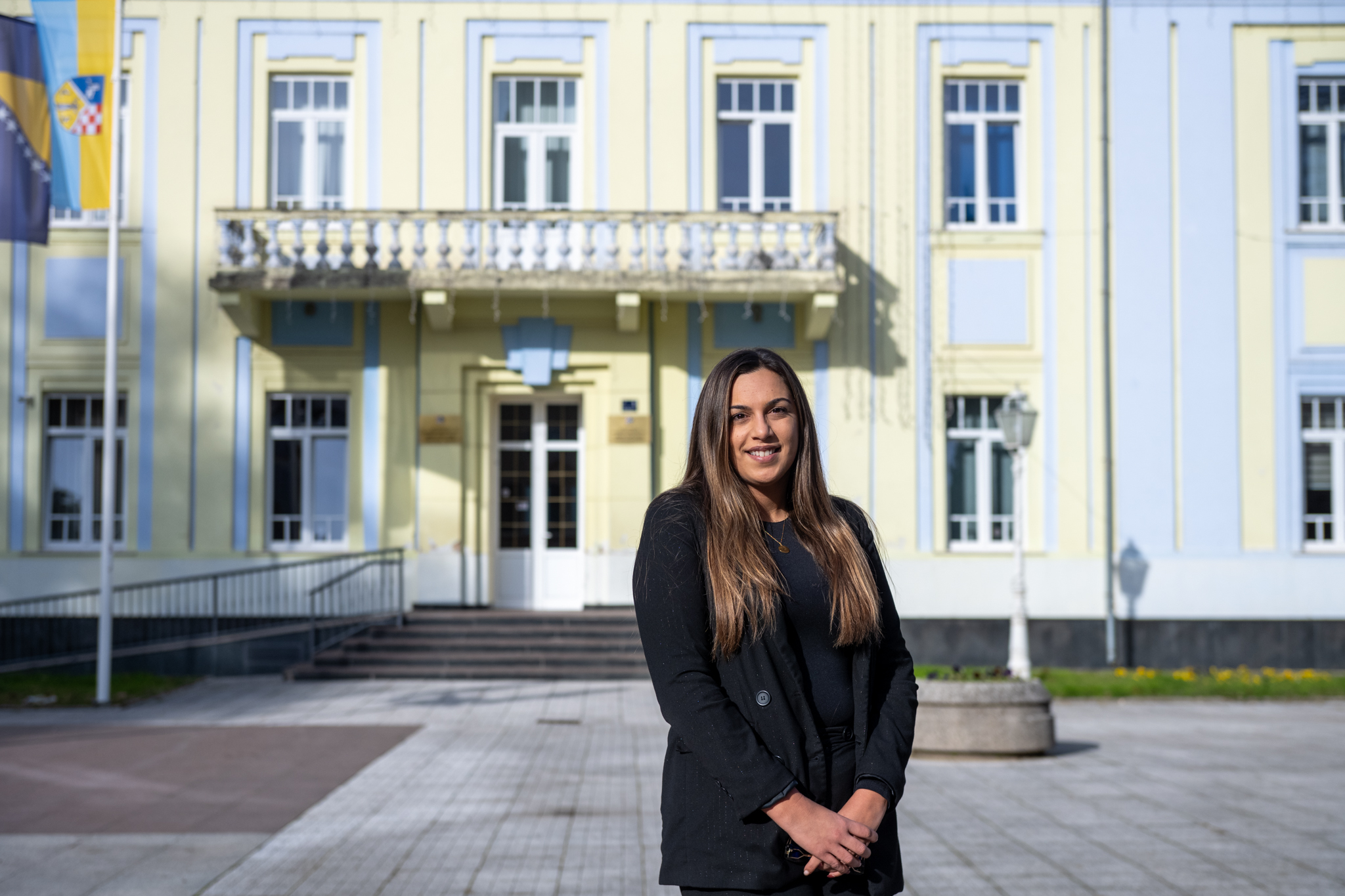 Woman with long hair in a black coat stands before a pale yellow building with white trim and balcony.