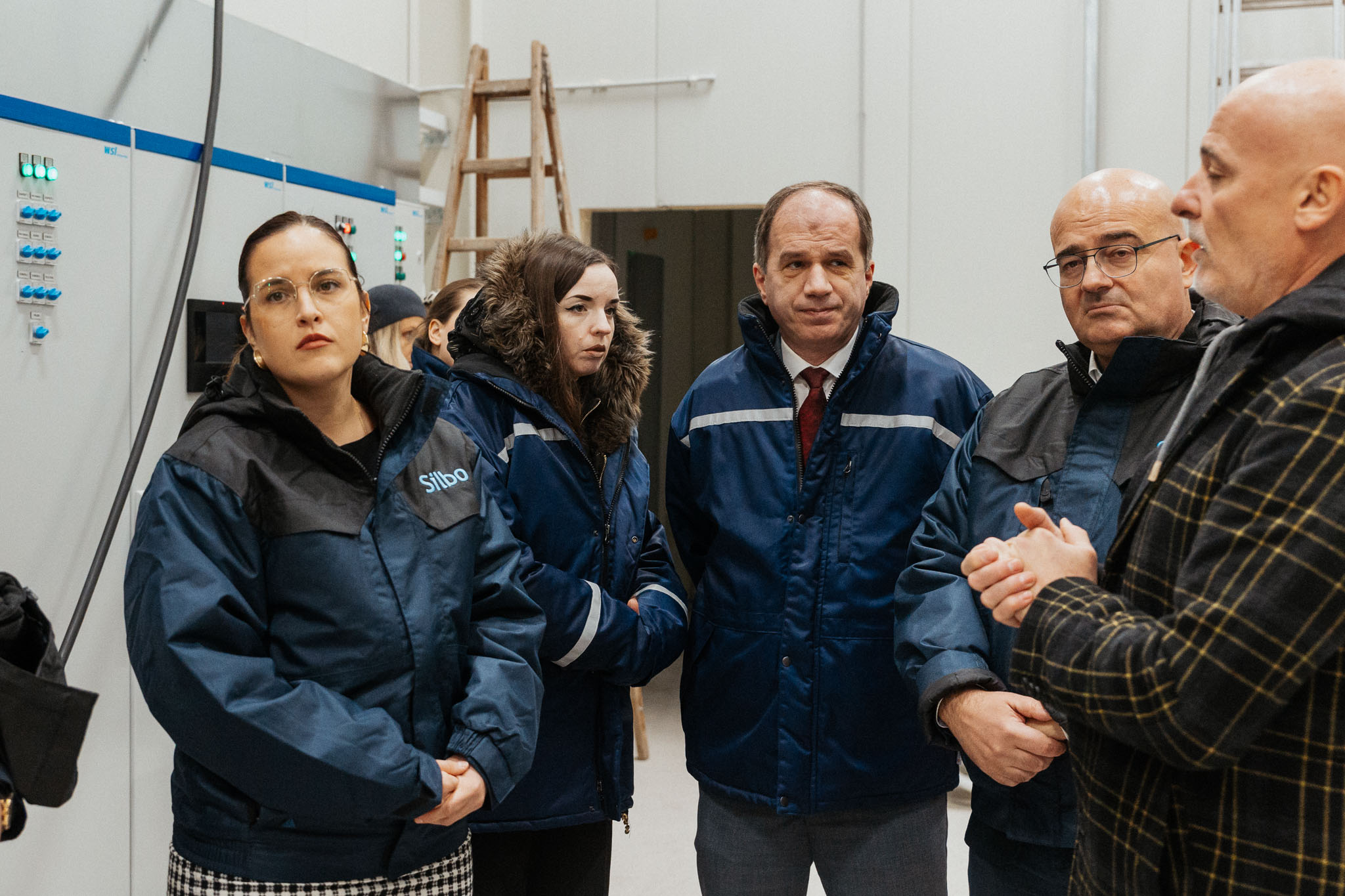 Small group in an industrial facility listens to a man in a blue jacket.