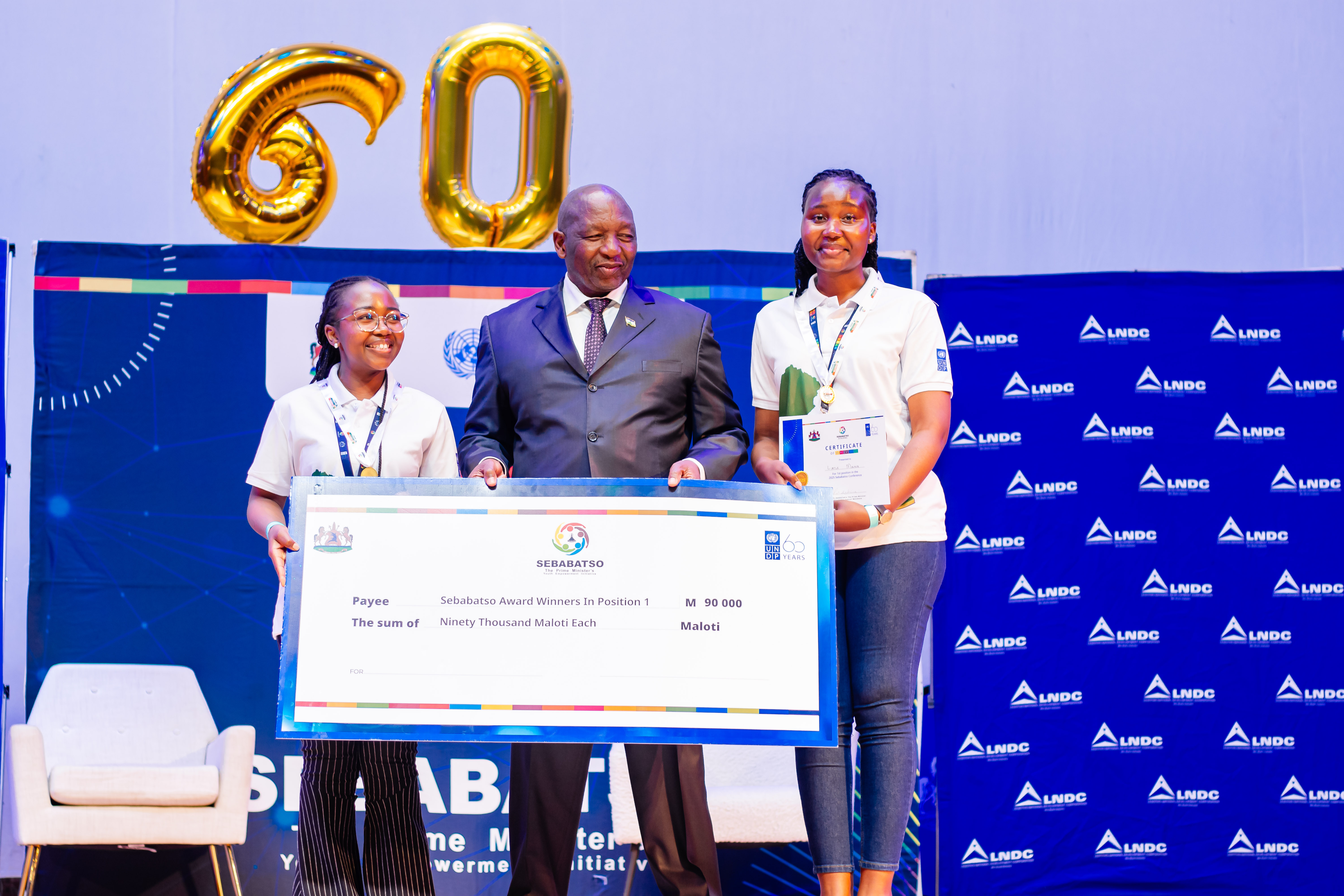 Group on stage holding an oversized check at a 60th anniversary event, with a blue backdrop.