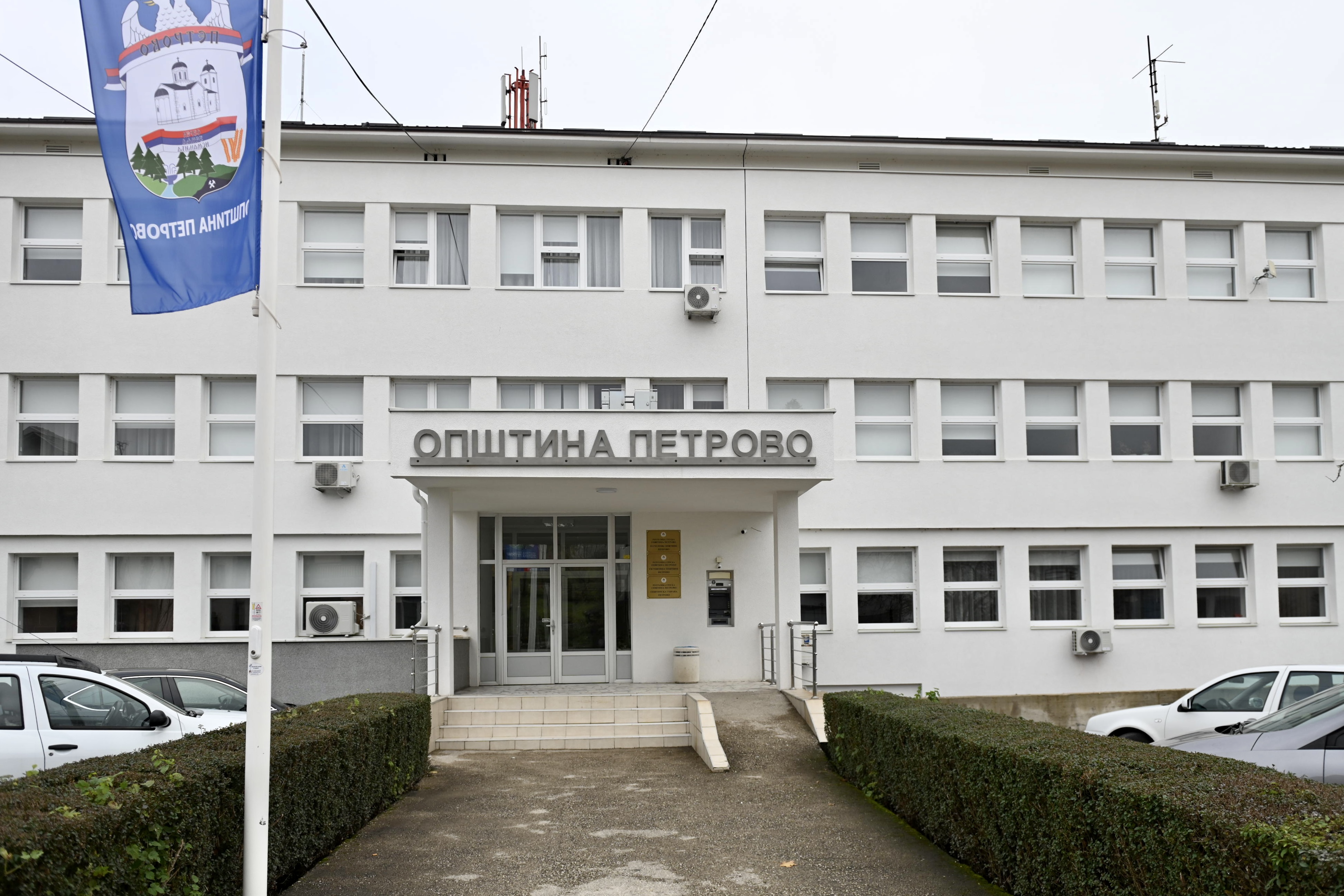 White, two-story building with central entrance; banner flag on left and two parked cars nearby.