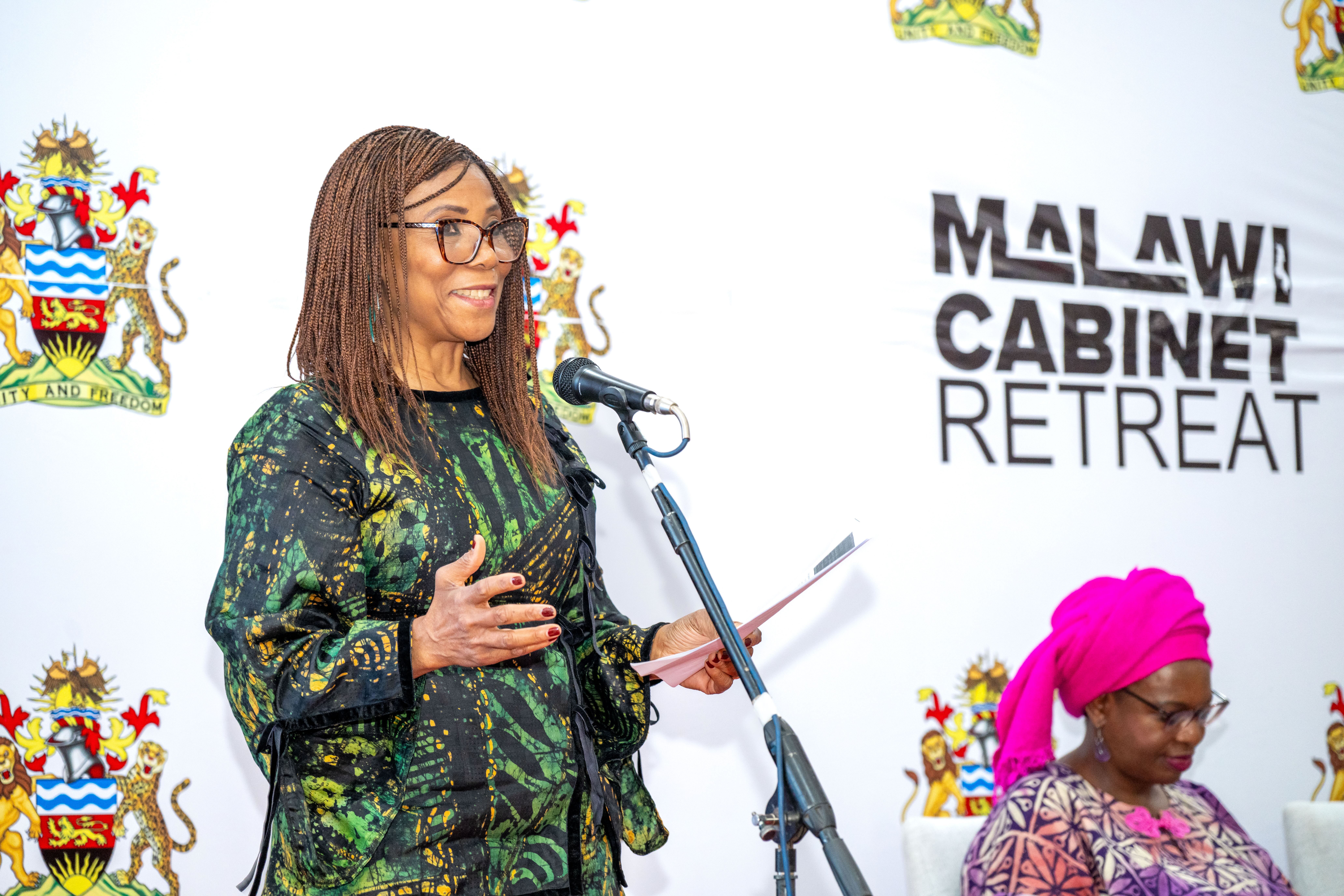 Speaker in green patterned dress at microphone during Malawi Cabinet Retreat; pink-haired attendee.