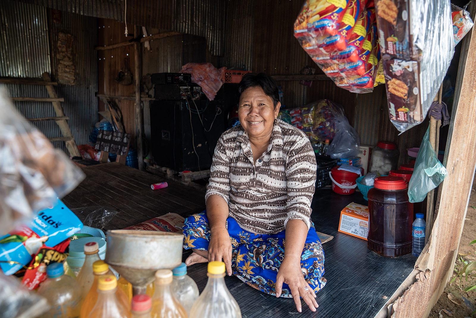 Market stall vendor in striped shirt sits amid jars, bottles, and bags.
