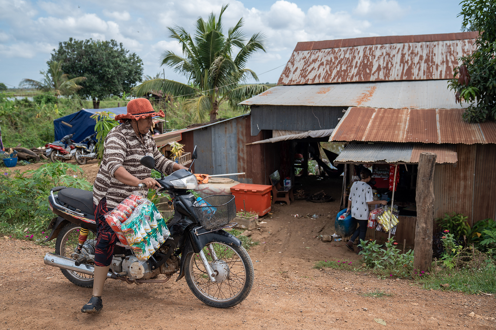 Woman on a motorcycle, wearing a prosthetic leg and holding bundled goods passes a rustic shop in a tropical village.