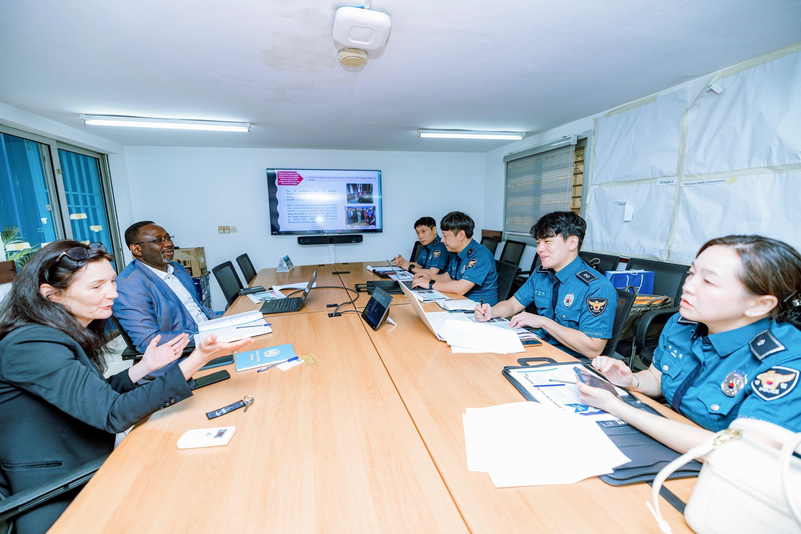 People around a long conference table in a bright room; several wear blue uniforms.