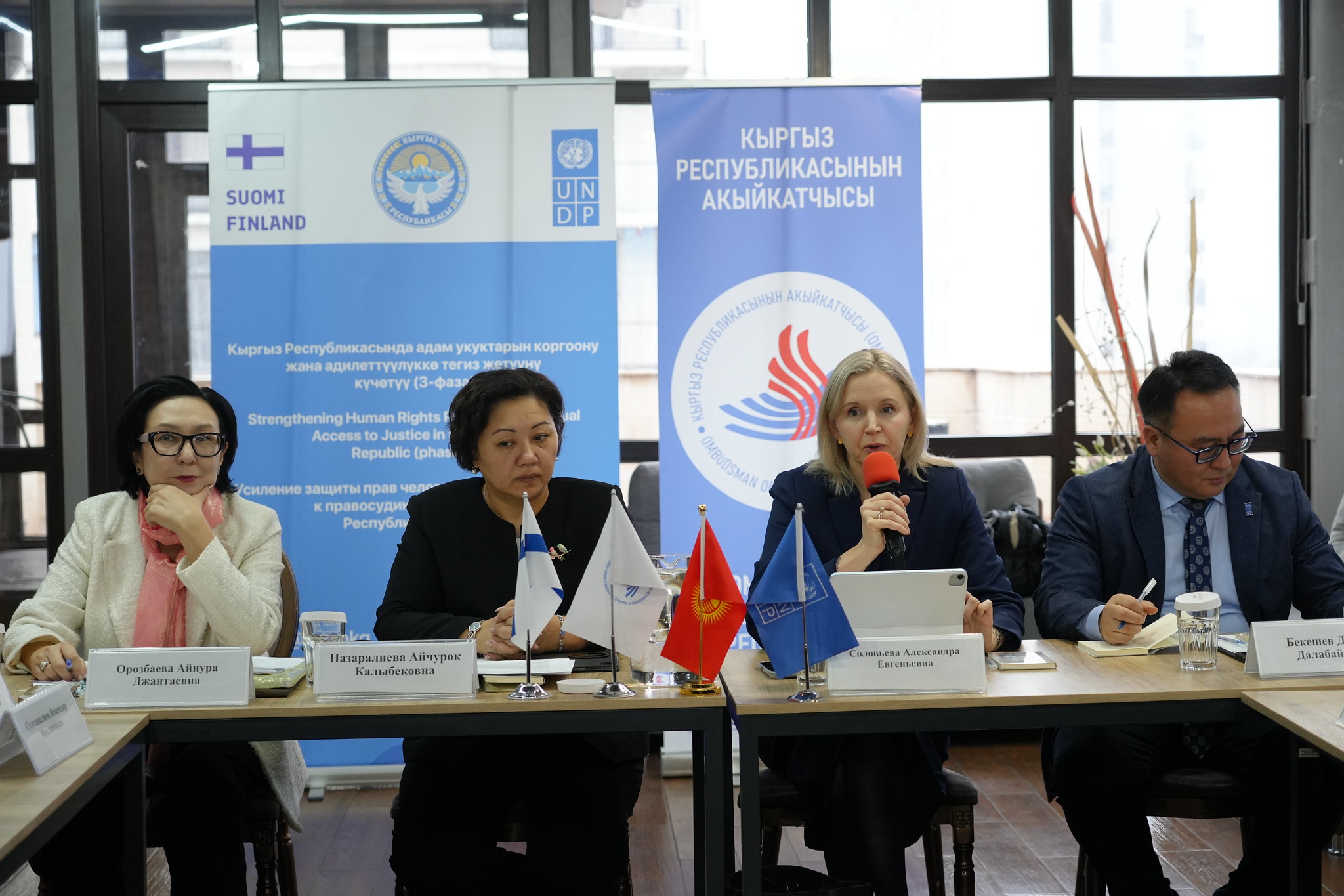 Panel of four speakers at a conference table, banners and flags behind.
