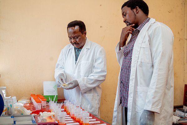 Two researchers in white lab coats stand by a table of cups and supplies in an orange-walled room.