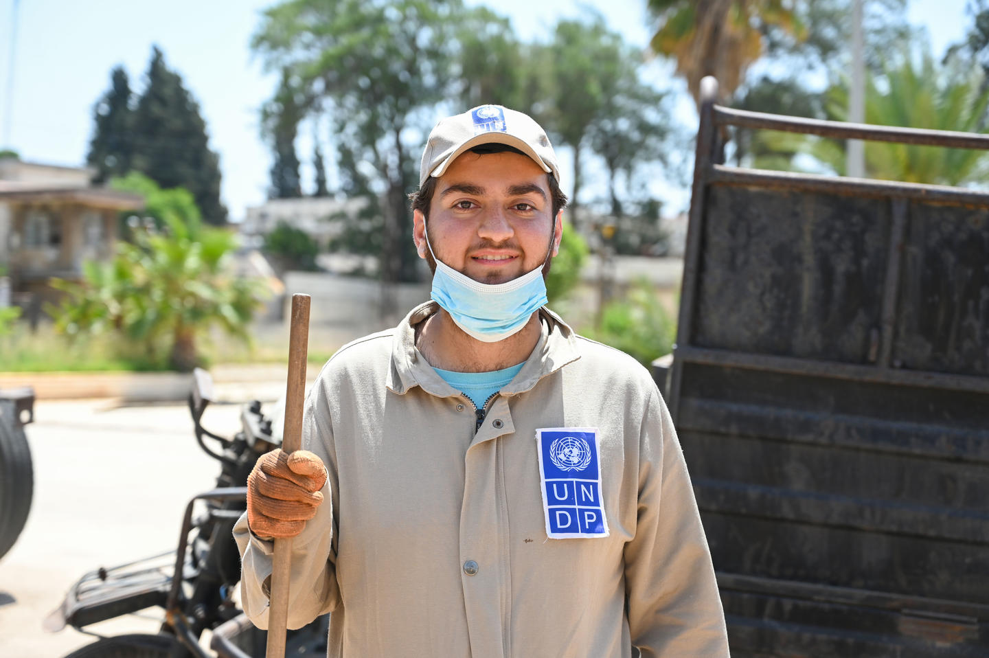 Man in beige shirt outdoors, blue mask around neck, holding a wooden handle near a truck.