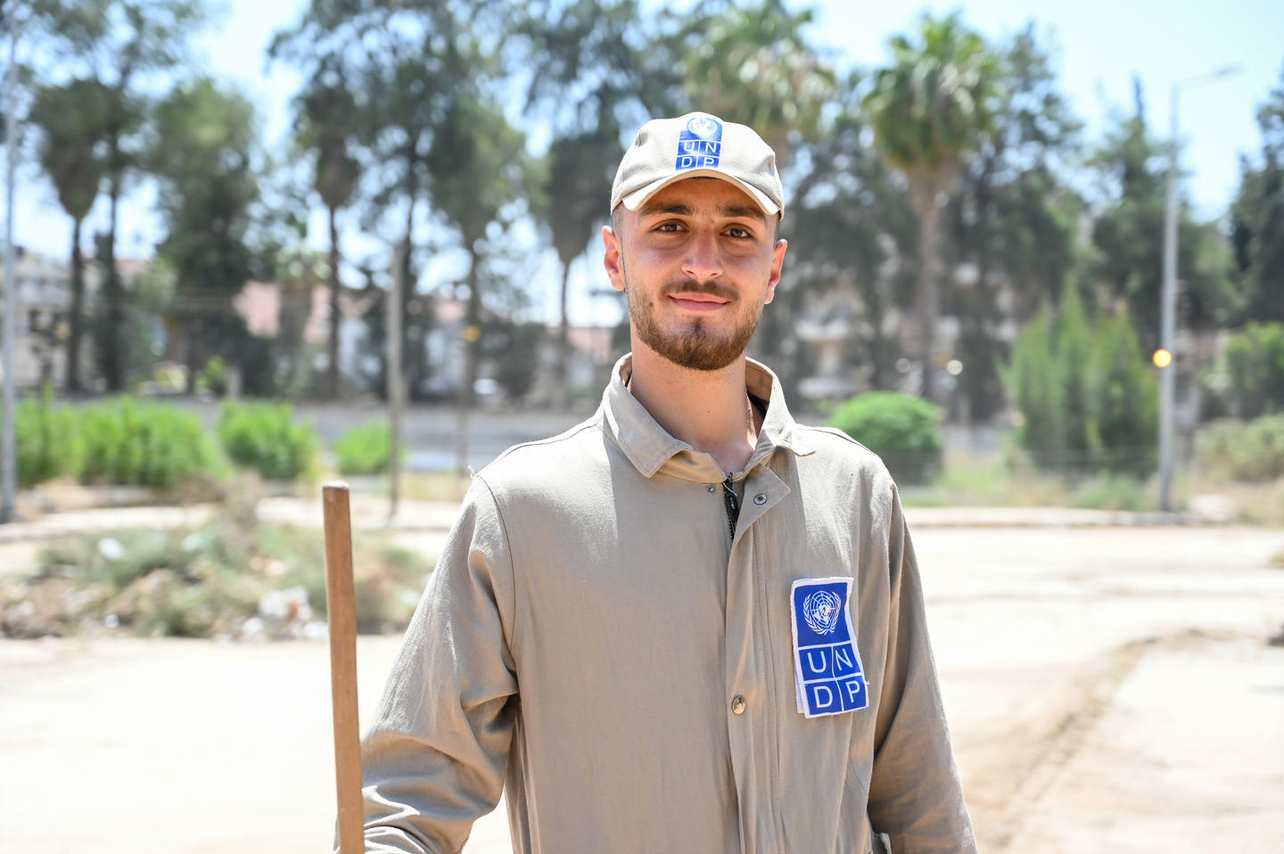 Person in beige uniform with blue badge on chest, standing outdoors near palm trees.