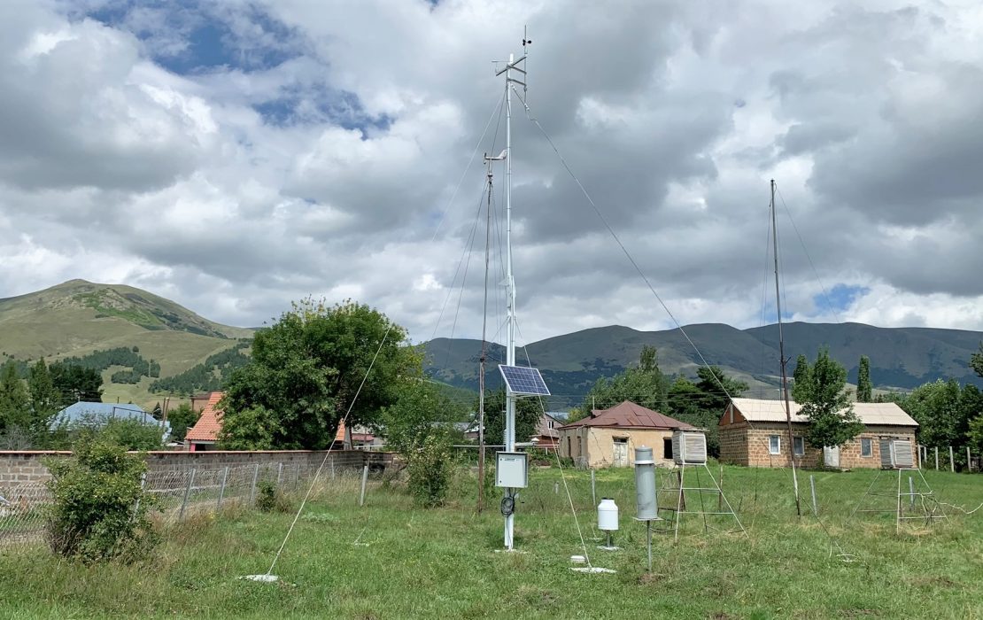Rural landscape with a tall metal antenna tower, small houses, hills, and a cloudy sky.