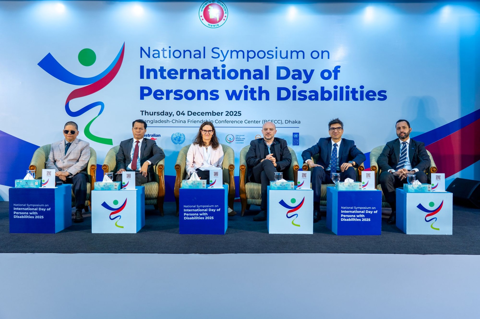 Photograph of panelists on stage at a disability symposium with blue banners.