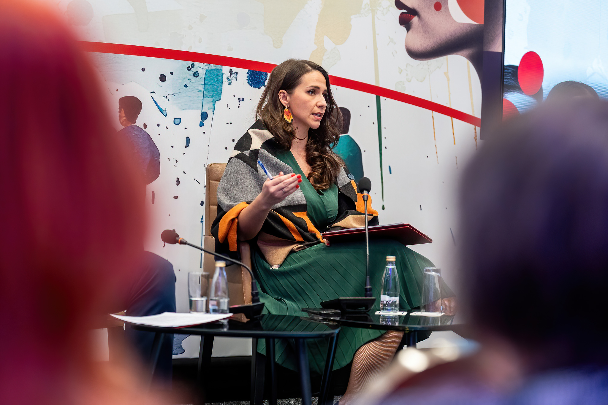 Speaker in green dress on stage, holding a notebook, addressing an audience.