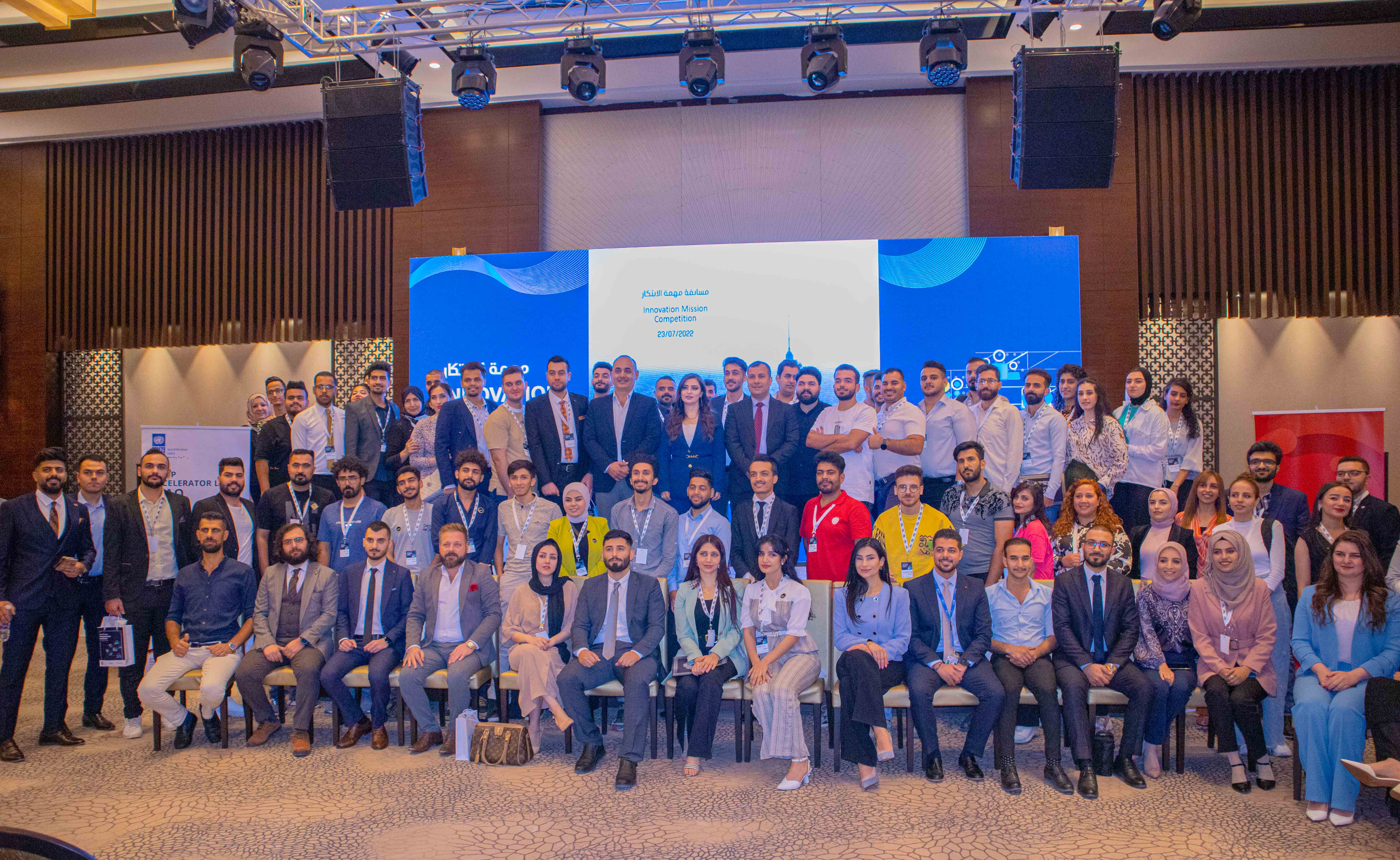 Group photo of a large crowd indoors on a conference stage with blue banners.