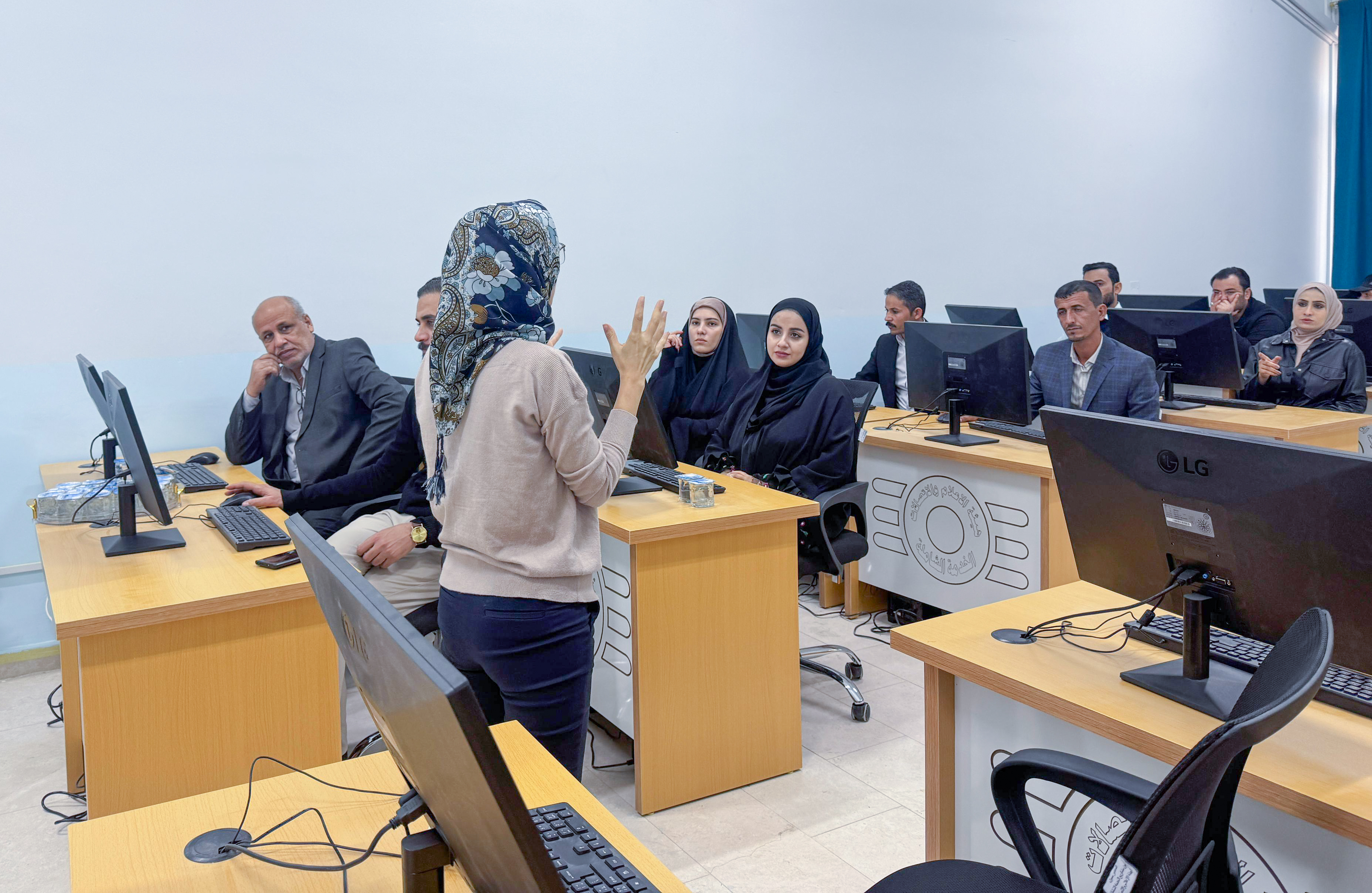 Bright computer lab; a presenter stands at a desk addressing a group at computers.
