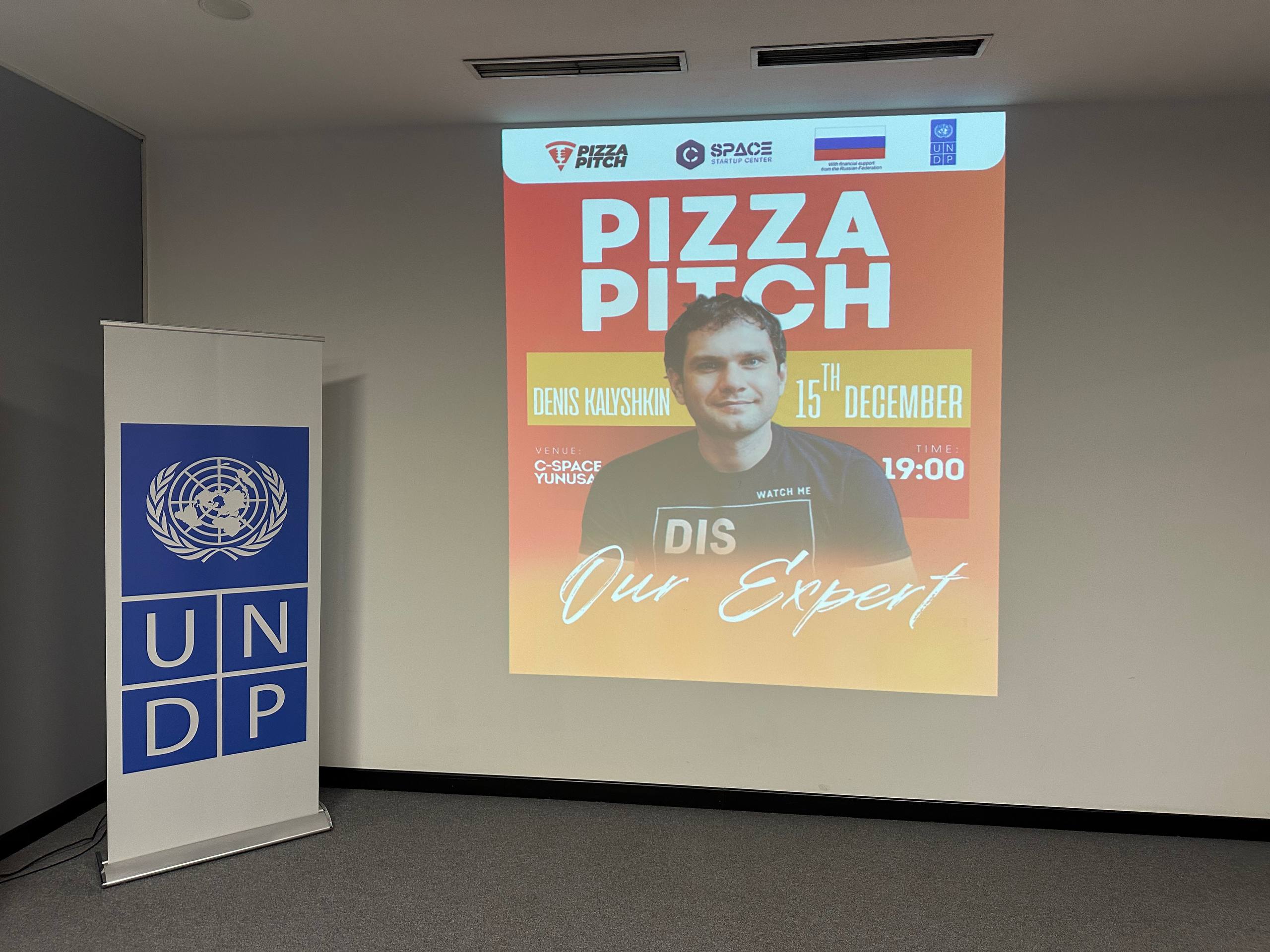 Conference room with an UNDP banner; projection displays a Pizza Night poster featuring a man.