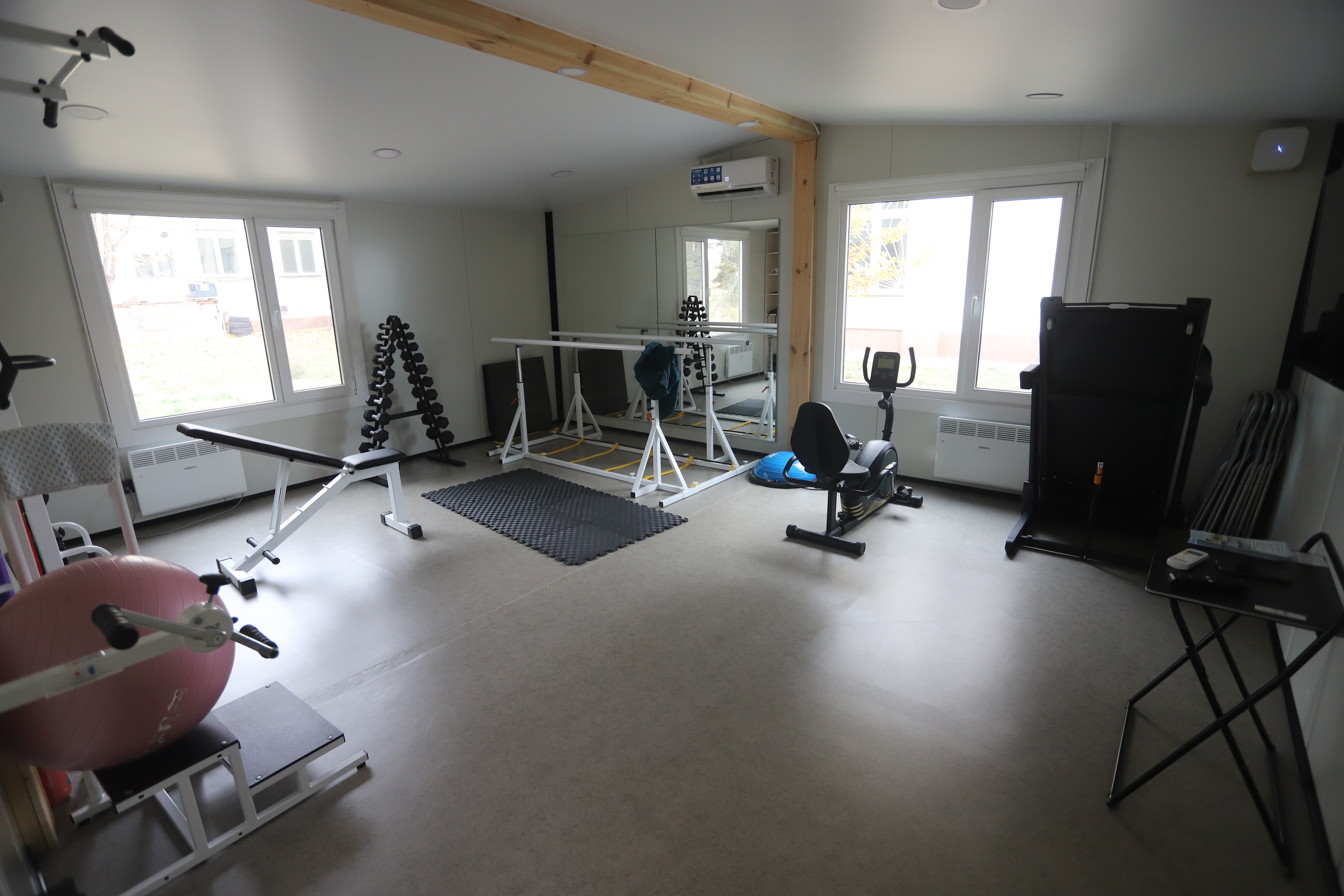 Bright rehabilitation gym with mats, treadmills, and equipment; a person stretches on a blue mat.