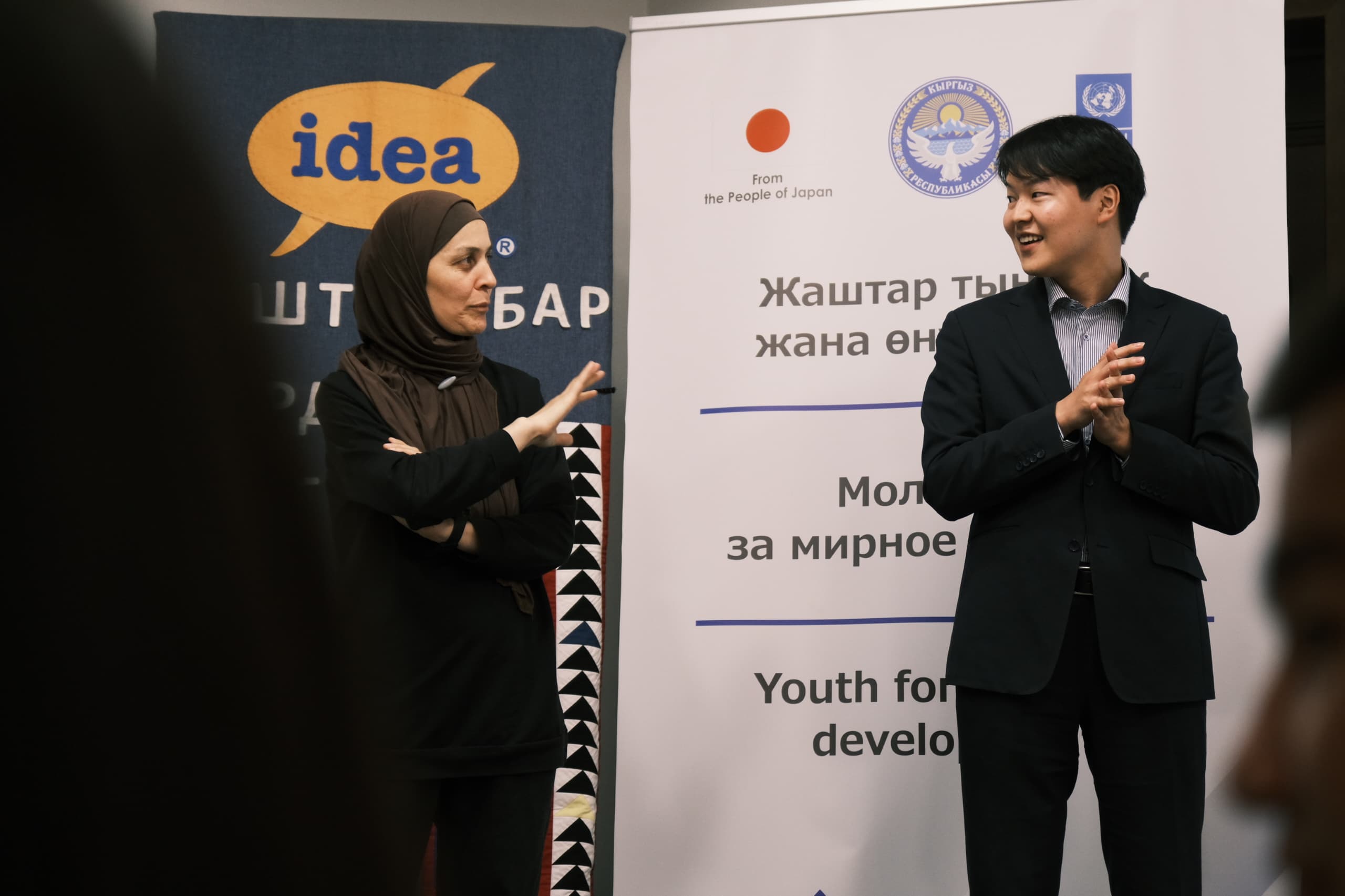 Two speakers on a stage with banners and logos; a woman in a hijab and a man in a suit.