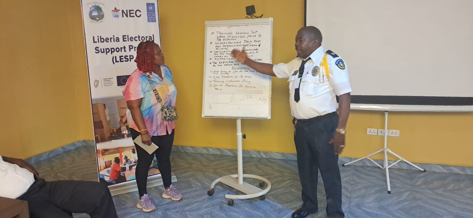 Photo: three people in a conference room; man with microphone near flip chart and banner.