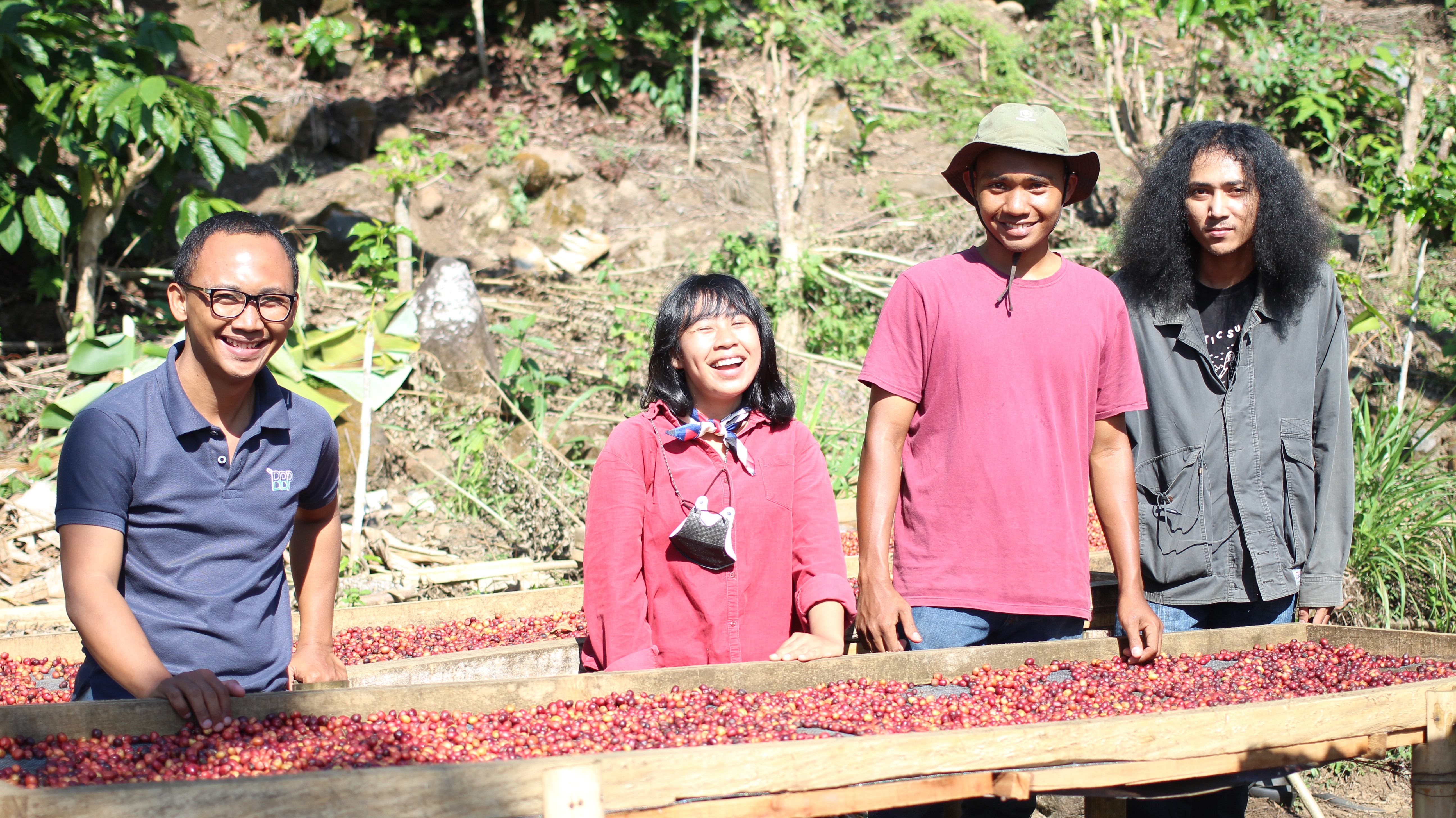 Five people smile behind a long wooden table outdoors, with coffee beans laid out